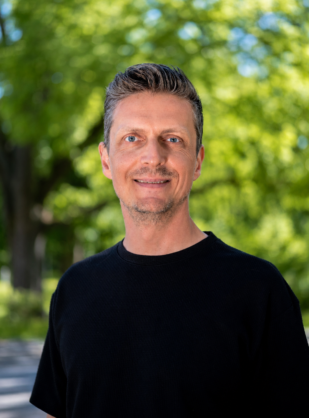 A middle-aged man with gray hair and blue eyes smiling outdoors, wearing a black shirt, with green trees and sunlight in the background.