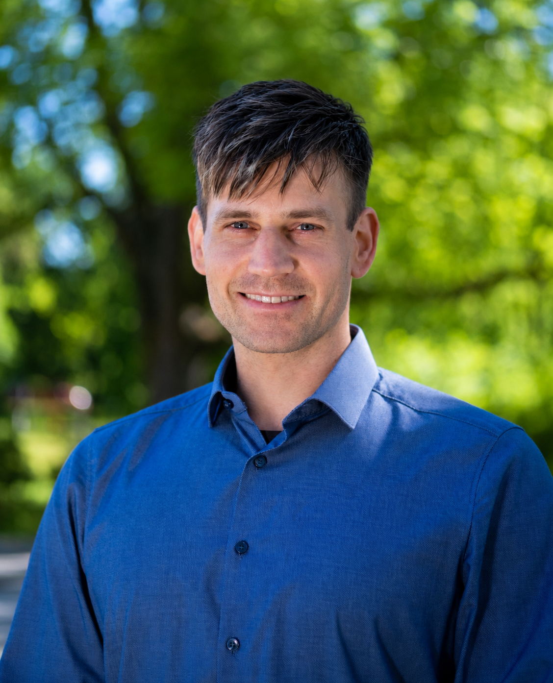 Portrait of a young man with dark hair and blue eyes, wearing a blue button-up shirt, smiling outdoors with green trees in the background.