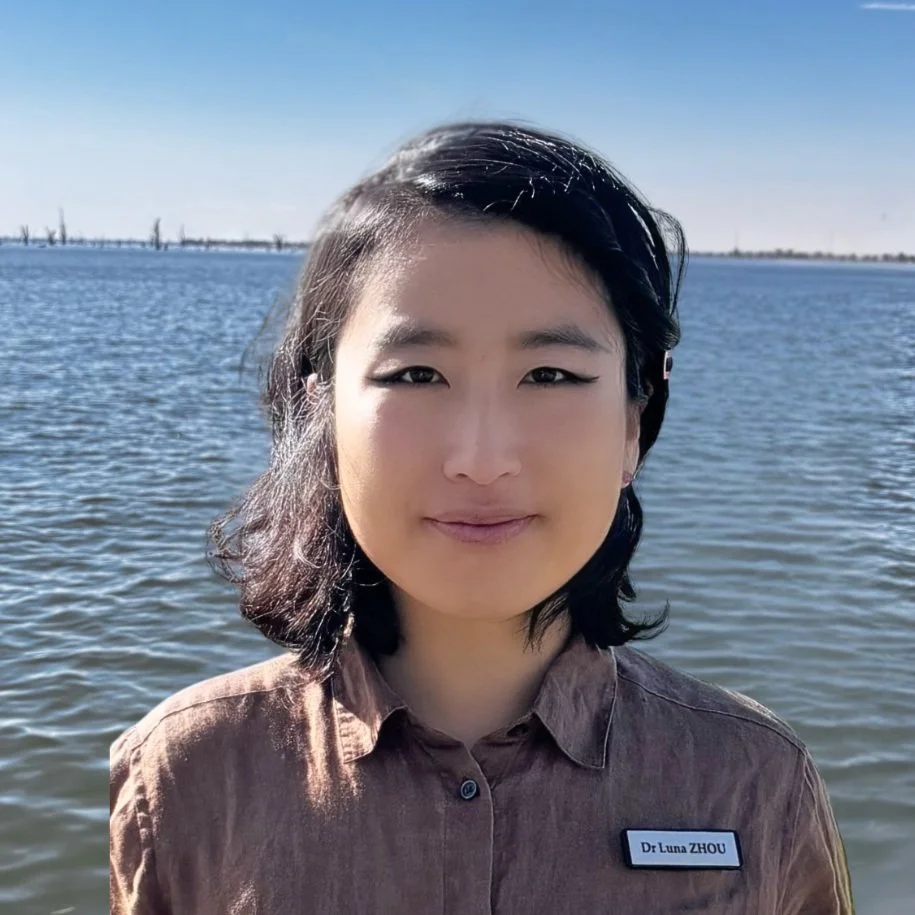 A woman with shoulder-length black hair, wearing a brown shirt with a name tag that says 'Dr. Luna ZHOU,' standing near water with a clear sky in the background.