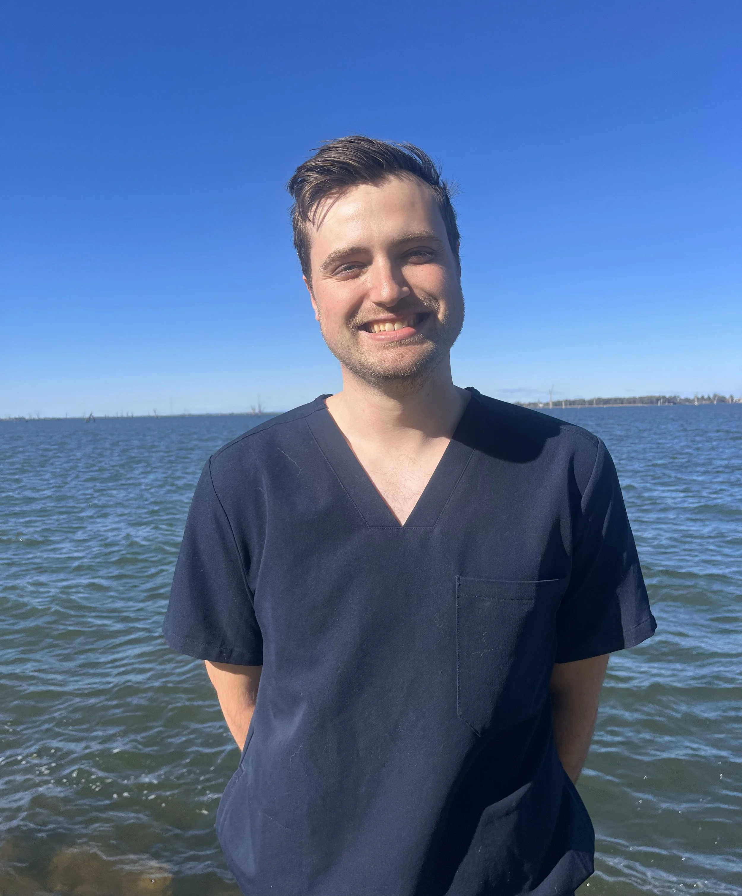 A young man with brown hair and a beard, smiling and wearing navy scrubs, standing near a body of water under a clear blue sky.