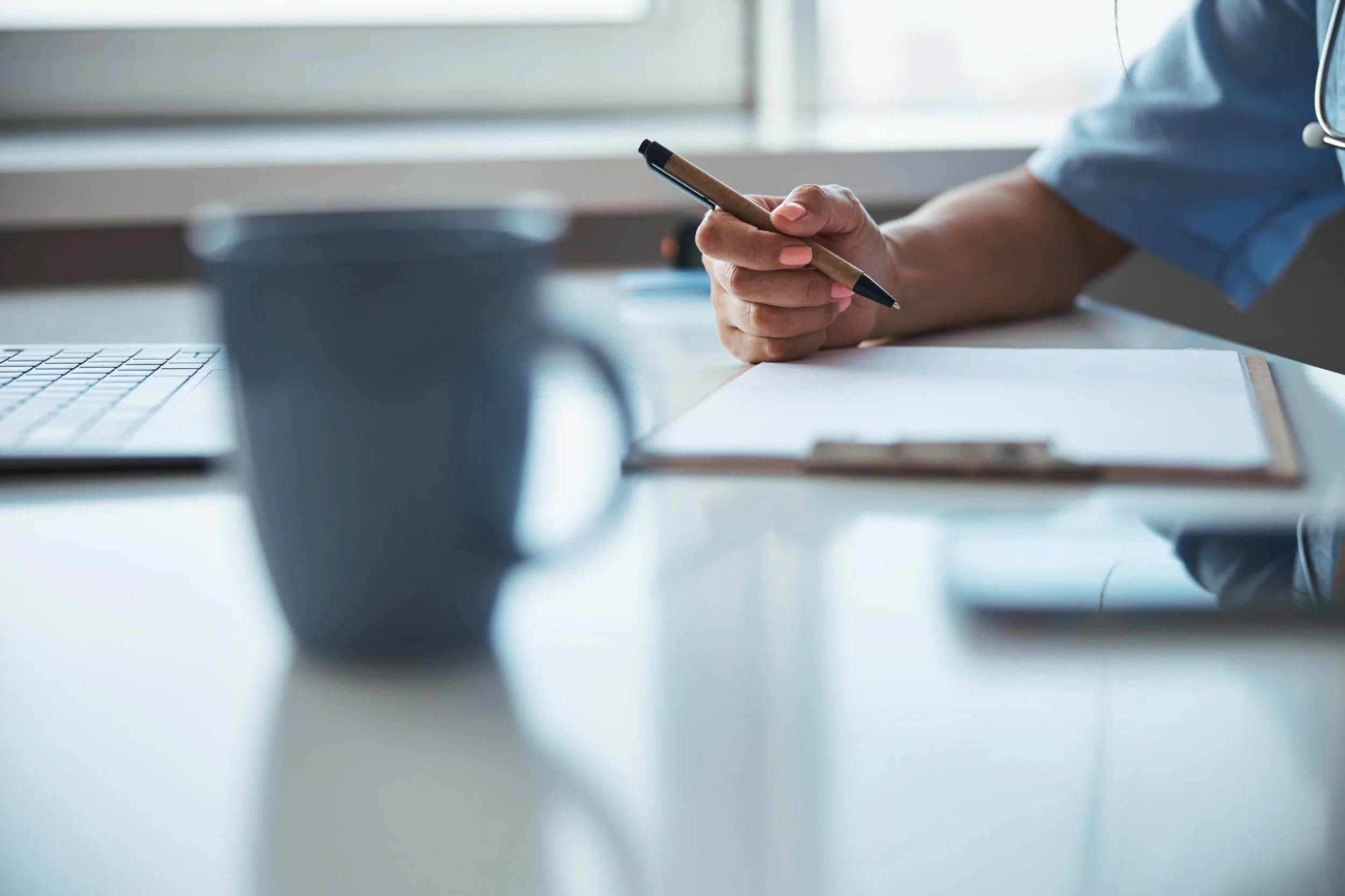A person in medical scrubs writing in a notebook on a clinic desk with a coffee mug and a laptop nearby.