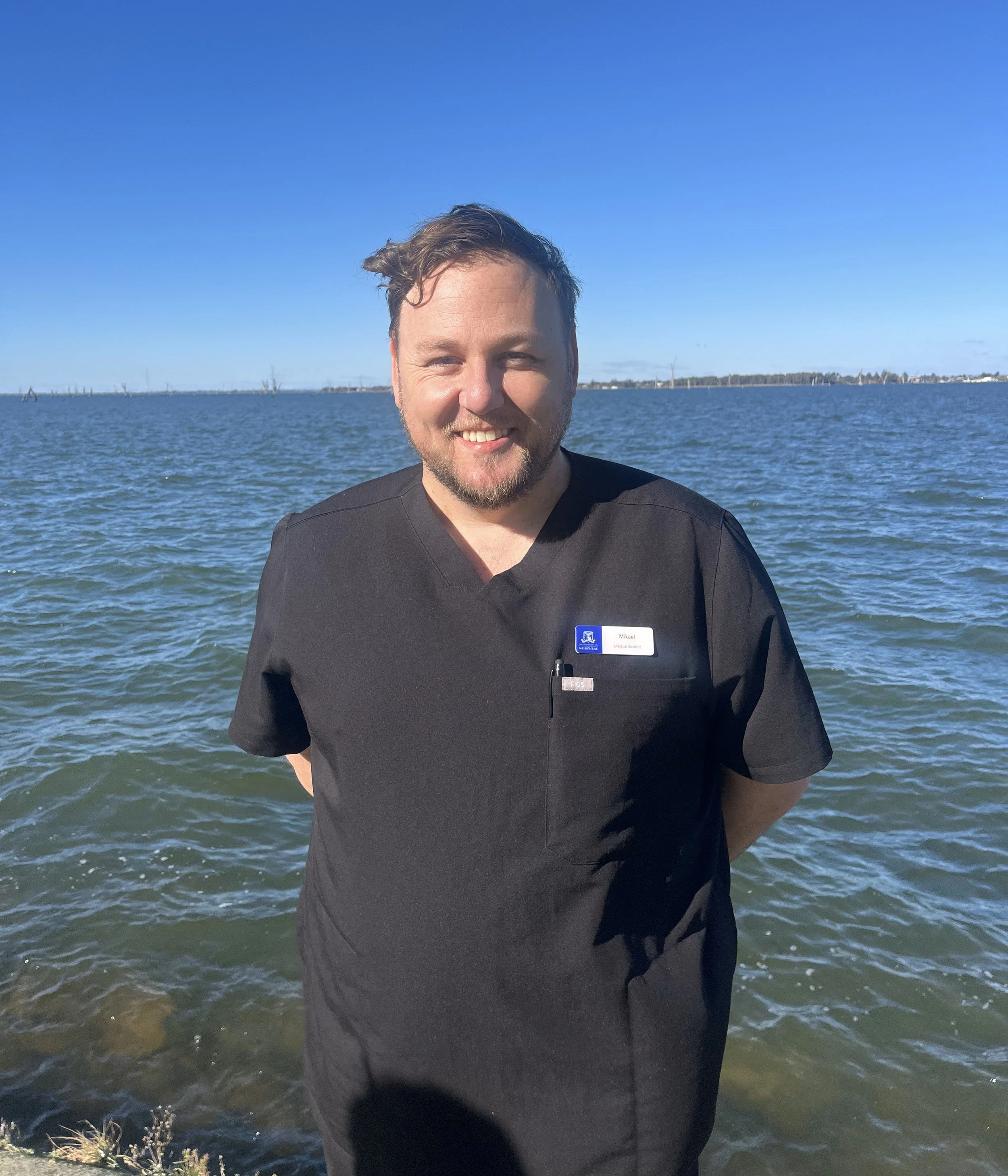 A smiling man in medical scrubs standing by a body of water on a sunny day.