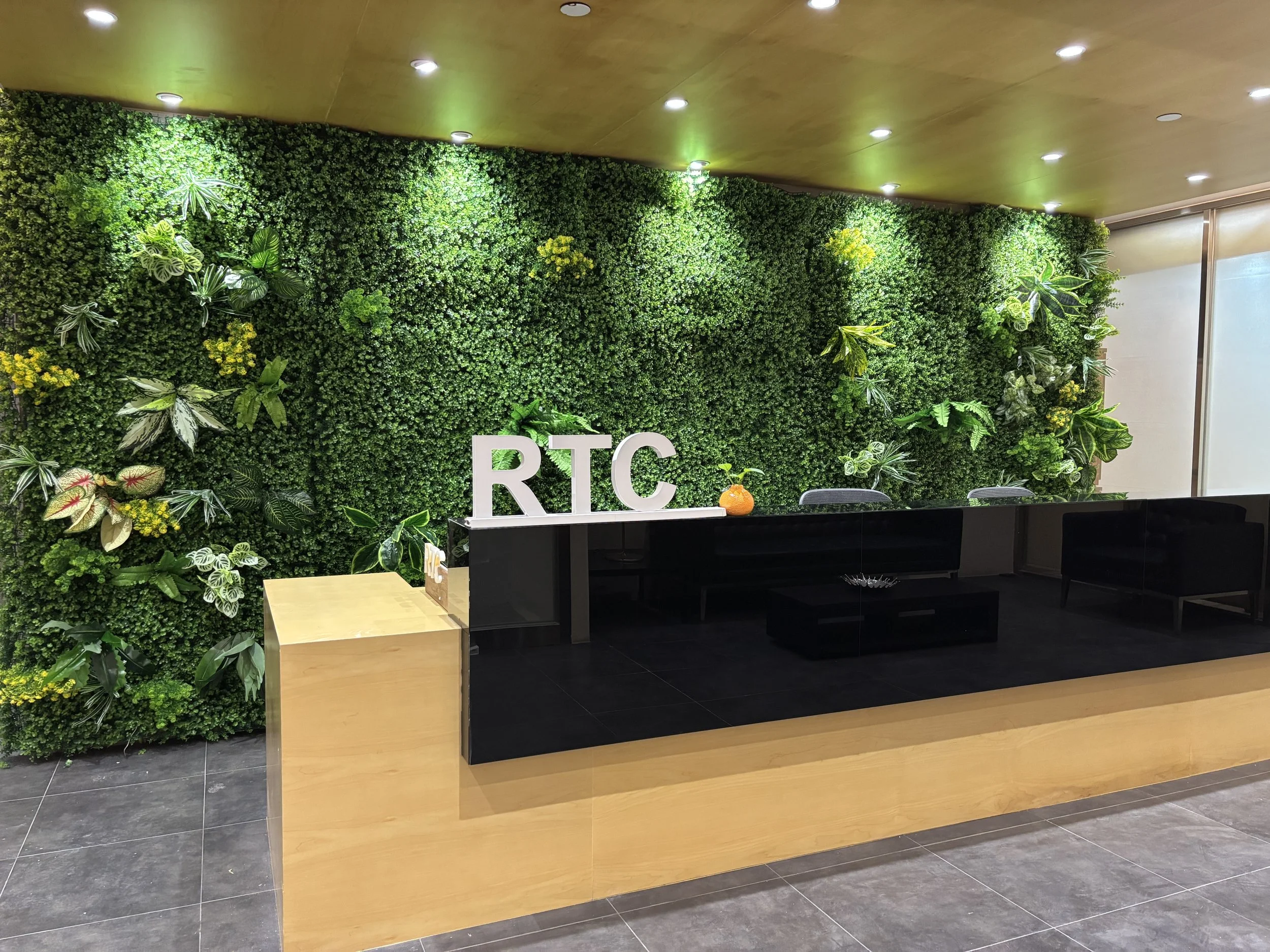 Modern office reception area with green wall decorated with plants and yellow flowers, black reception desk with white 'RTC' letters and a small pumpkin decoration, and wooden accents.