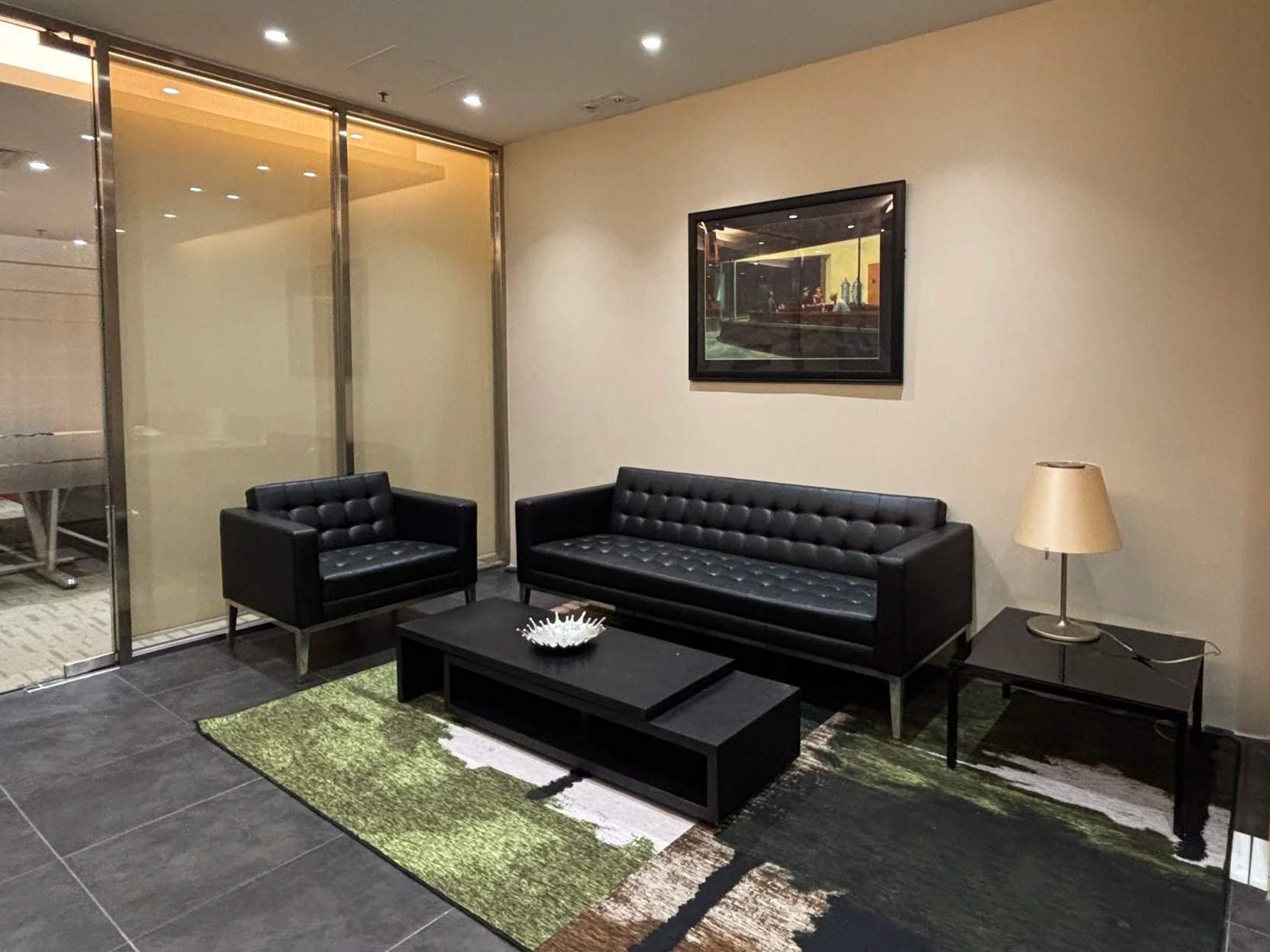 Modern hotel lobby sitting area with black leather furniture including a sofa and an armchair, a black coffee table with a decorative bowl, a side table with a lamp, and a framed artwork on the wall. There is a glass partition to the left and a patte