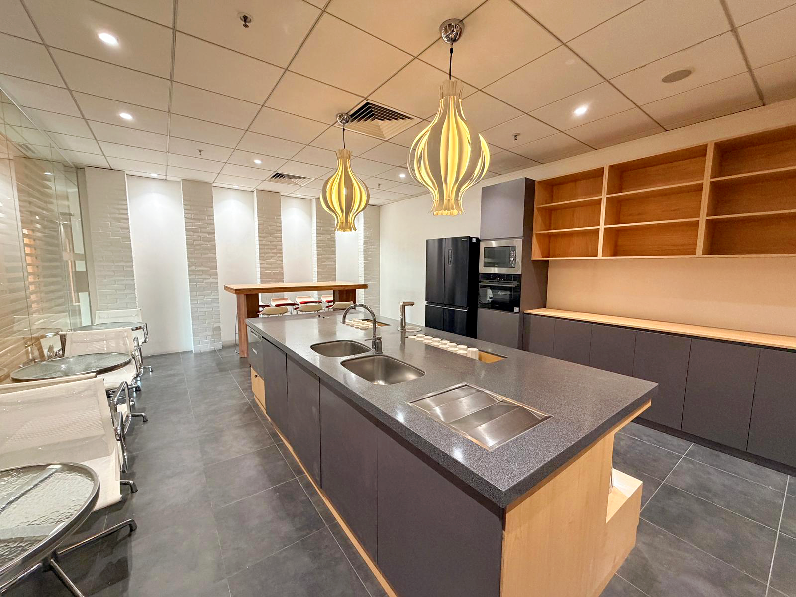 Modern kitchen with black and wooden cabinets, a large kitchen island with double sinks, two hanging pendant lights, an open shelf on the wall, a refrigerator, a microwave, and a bar-height table with chairs in the background.