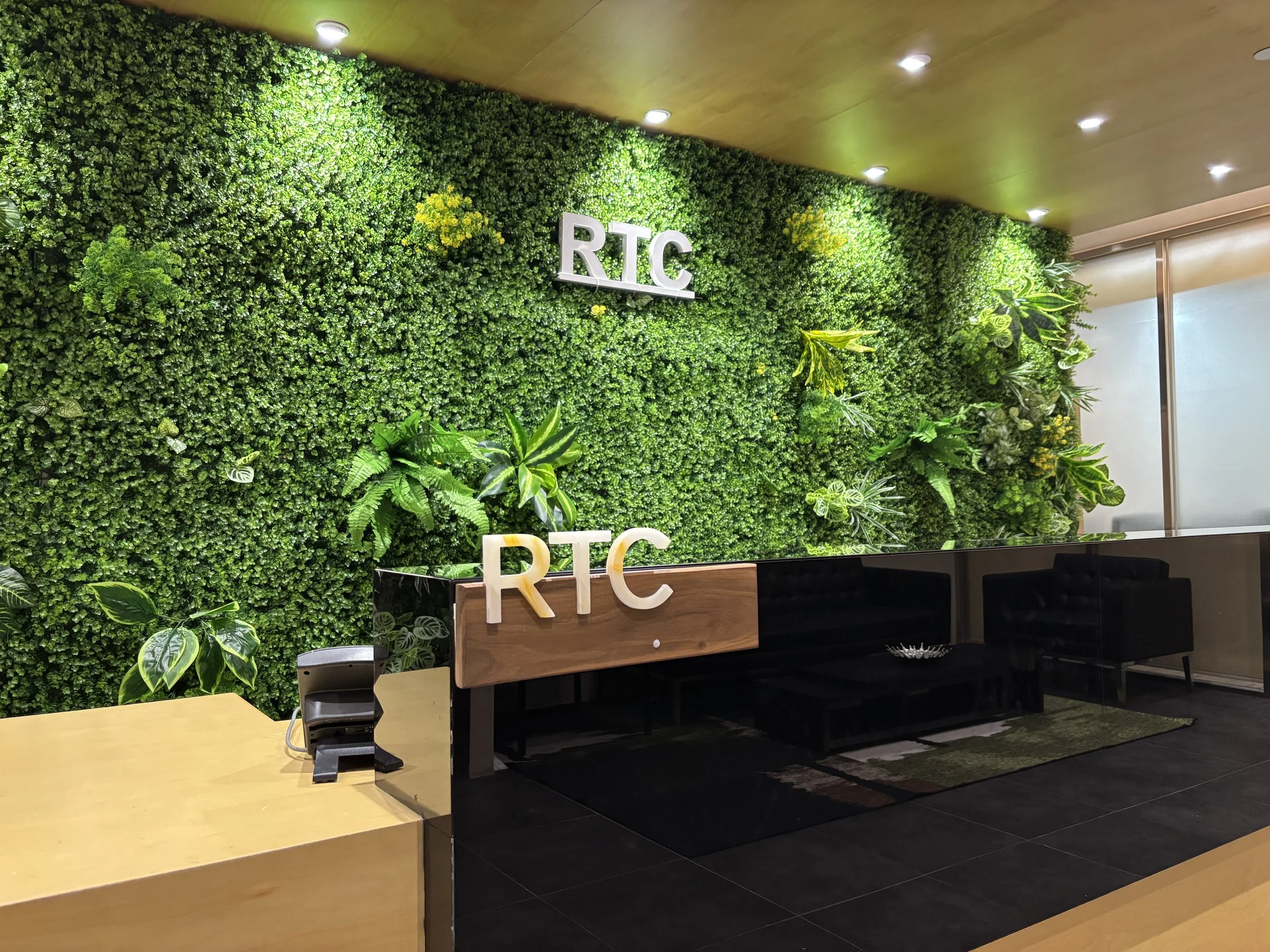 Reception desk area with a green plant wall in the background and two cut-out letters 'RTC' in white on the greenery. The desk is black with a wooden accent and has the letters 'RTC' on it. There are some orchids and leafy plants on the wall and near