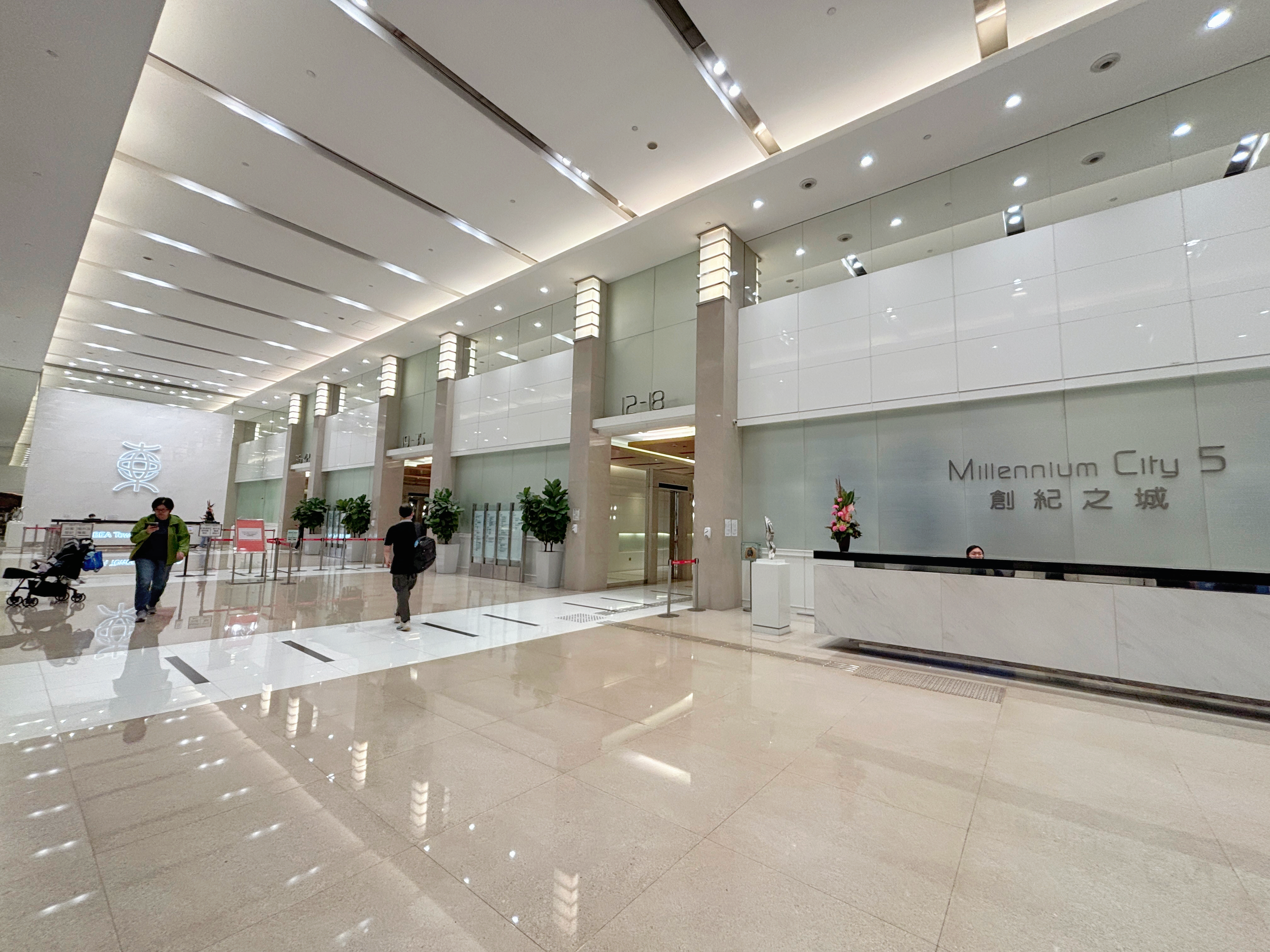 Interior of Millennium City 5 shopping mall with a reception desk, name sign, plants, and people walking.