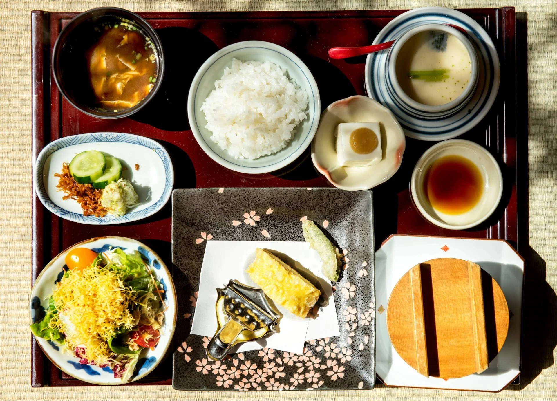 A traditional Japanese meal served on a tray, featuring rice, miso soup, tempura, pickled vegetables, tofu, and various side dishes.
