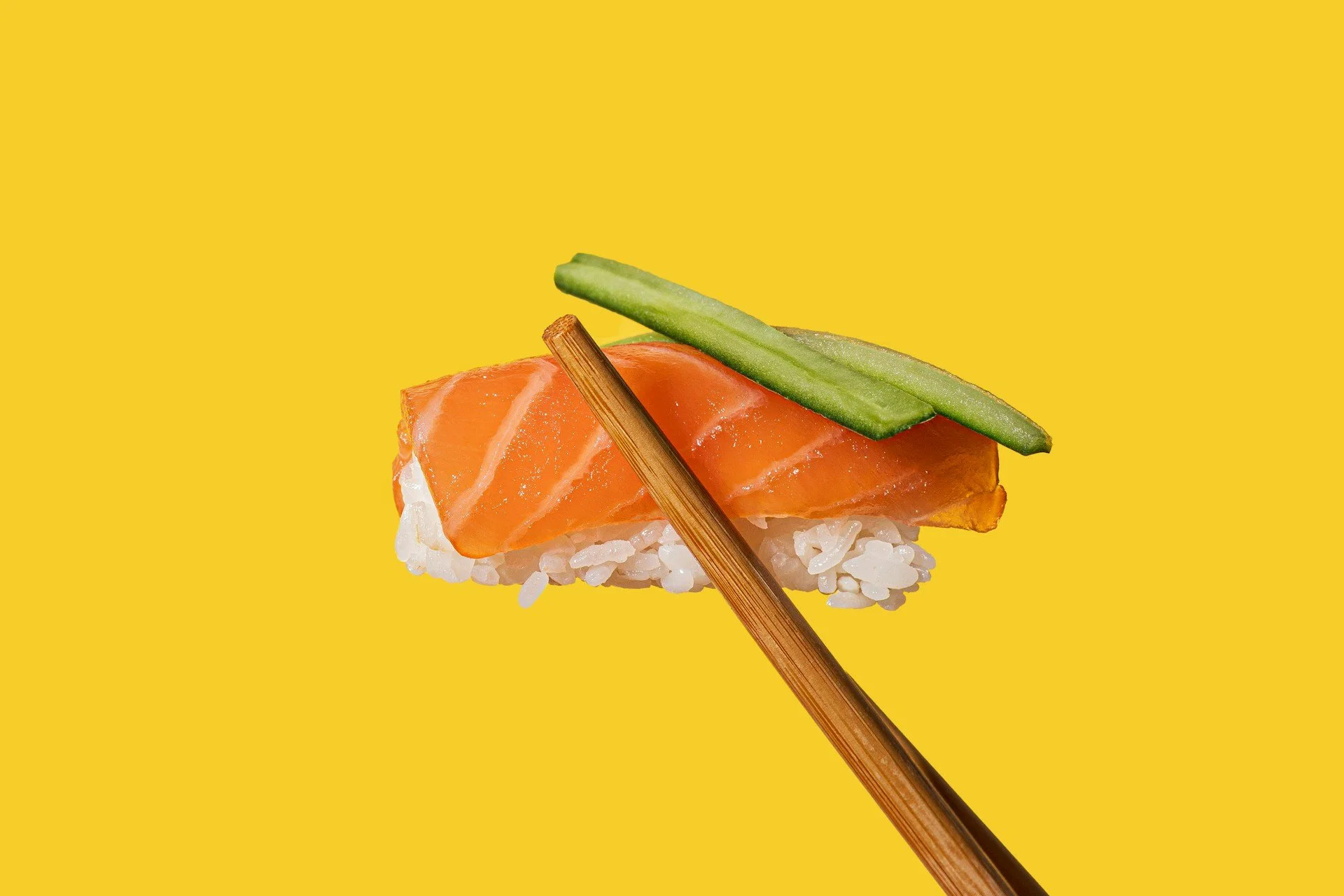 Chopsticks holding a single piece of sushi (rice, salmon, and cucumber) over a yellow background.