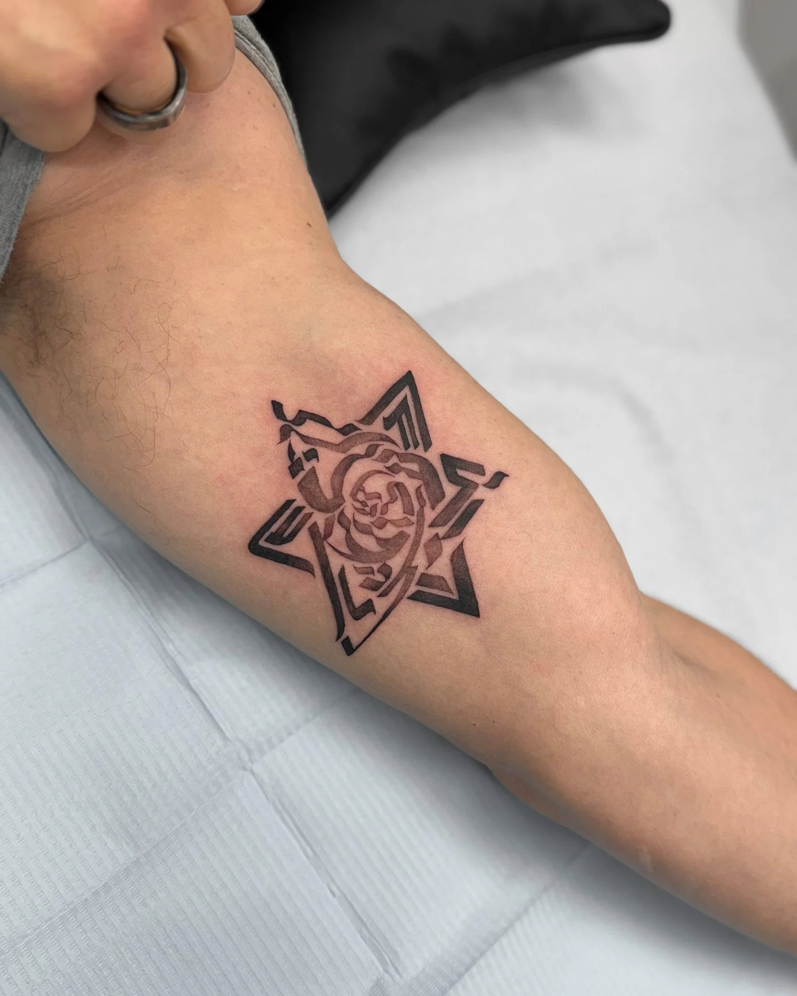A close-up of a person's arm with a geometric star-shaped tattoo featuring a lion's face at the center. The arm has some fine hair and light skin tone. The person is wearing a silver ring on their finger, which is partially visible, and is sitting on a white textured surface.