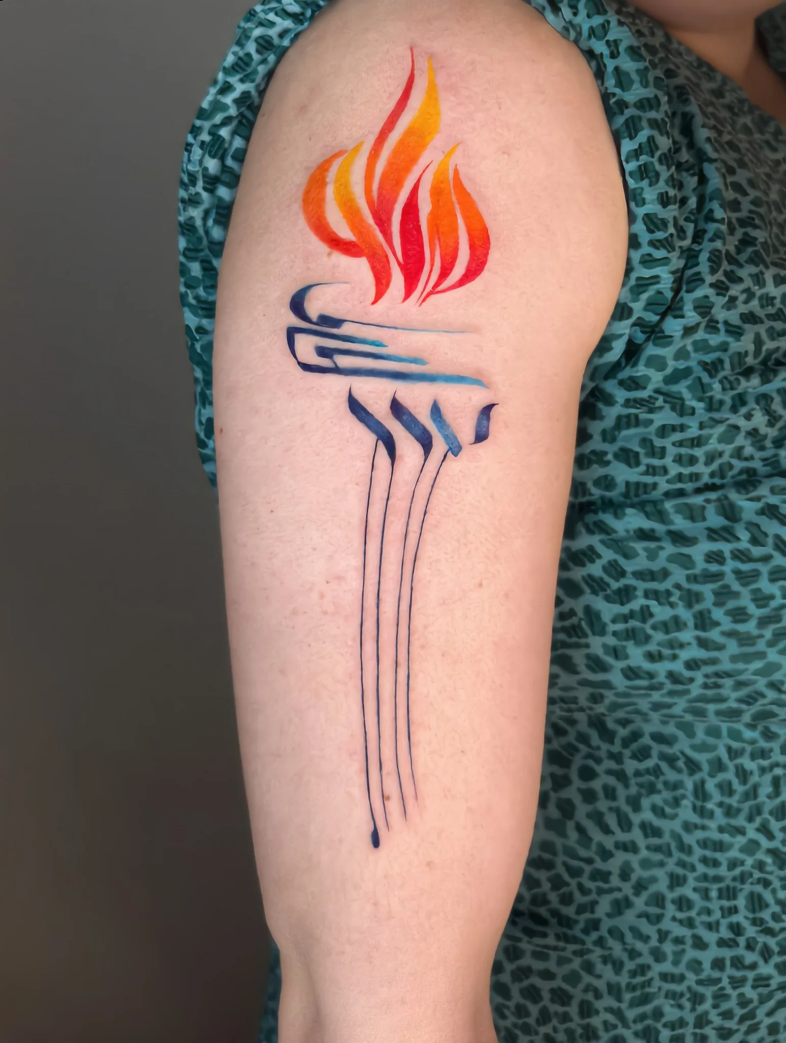 Colorful tattoo of a flame with red, orange, yellow, and blue hues, located on a person's arm.