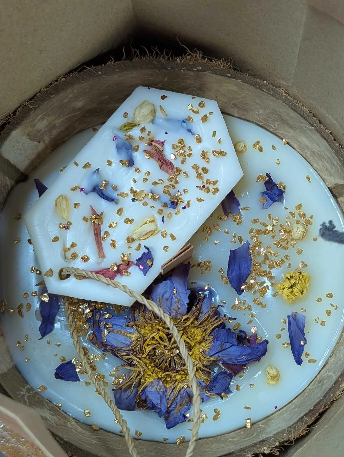 A round, pale blue candle with embedded dried flower petals and surrounded by scattered dried flower petals and gold flakes.