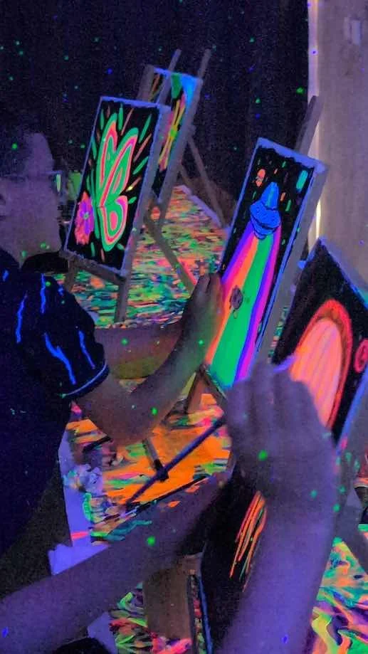 People painting glow-in-the-dark artwork in a dark room illuminated with blacklight, with colorful, vibrant, and glowing paint on canvases and tables.