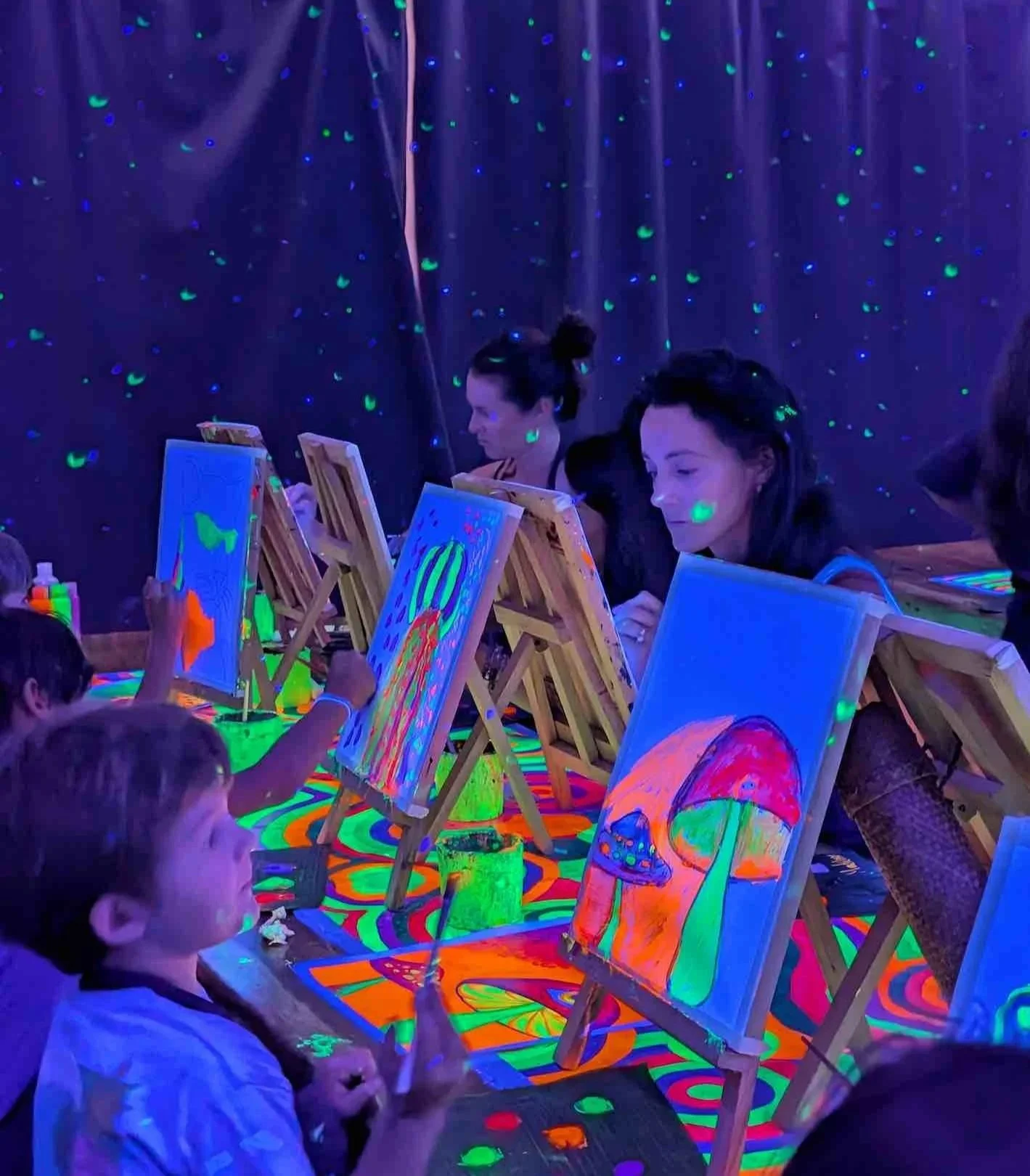 A young girl with face paint under blacklight, painting on a canvas as colorful glow-in-the-dark paint appears in the dark. There is a water bottle on the table.
