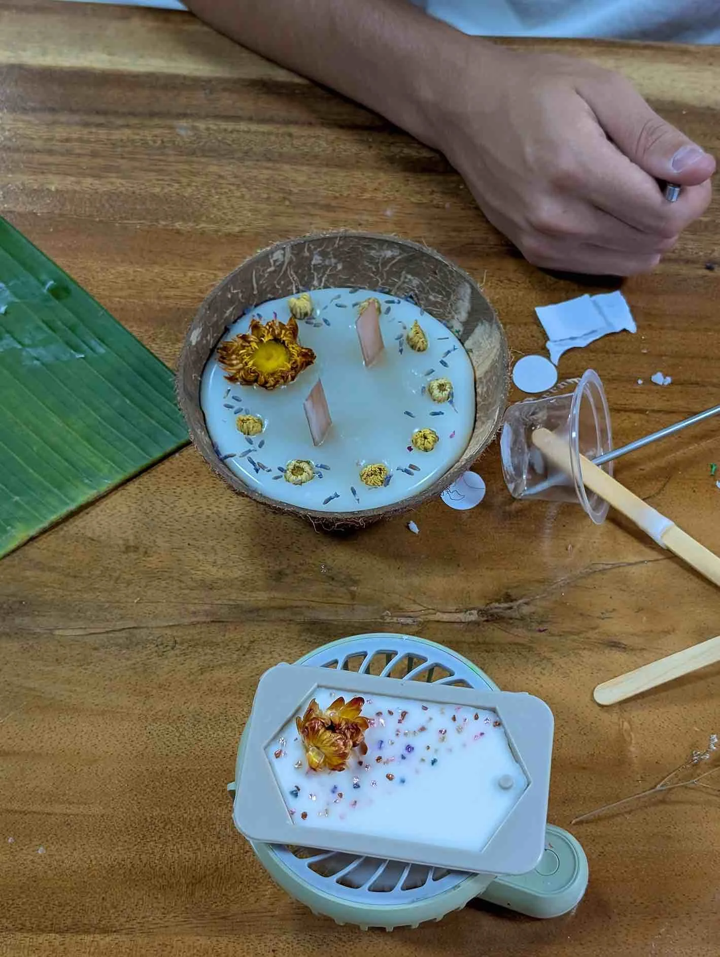 A carved coconut shell contains a white candle with dried flowers and small purple flowers floating on the surface. In front, there is a white wax block with dried flowers and colorful sprinkles, resting on a small fan.