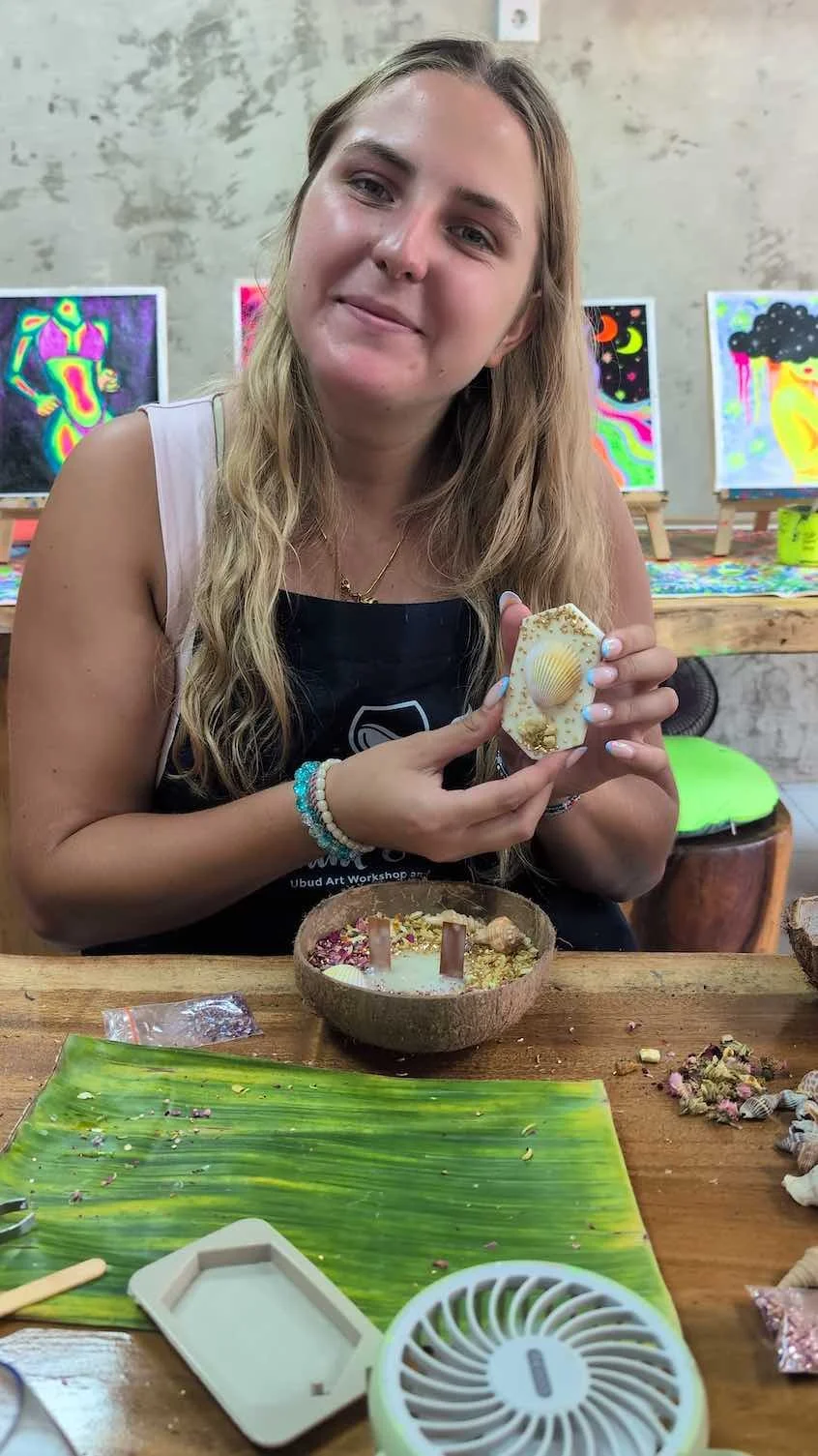 A young woman with long blonde hair holding a decorated soap with a shell in the center. She is sitting at a wooden table with art supplies and a bowl of soap scraps in front of her. Behind her, colorful art pieces are displayed on small easels.