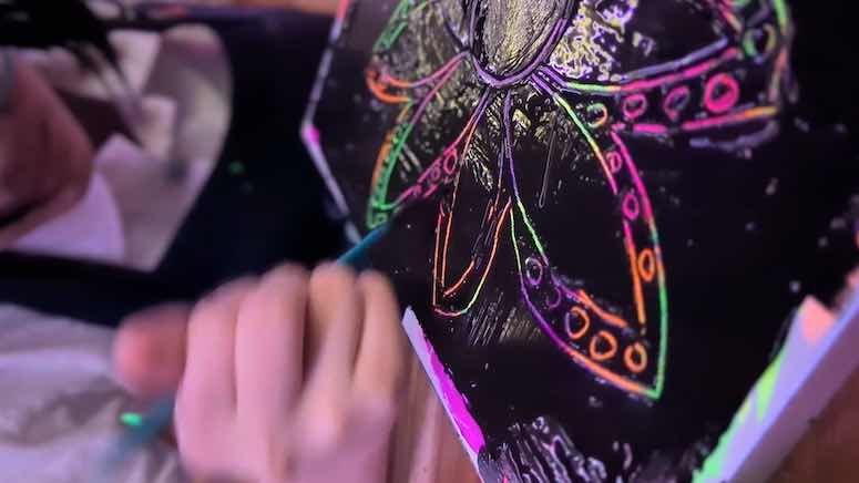 Child drawing a colorful neon butterfly on black paper with a glow-in-the-dark art set
