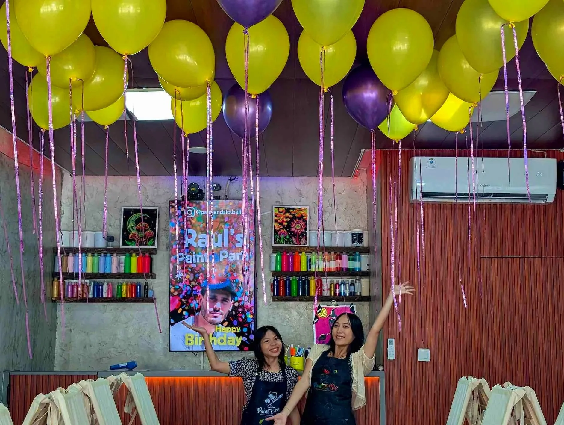 Celebration at a paint party with yellow and purple balloons on the ceiling, two women smiling and holding a sign that says 'Raul's Paint & Sip Happy Birthday', shelves of colorful paint bottles, and a digital birthday message on a screen in the background.