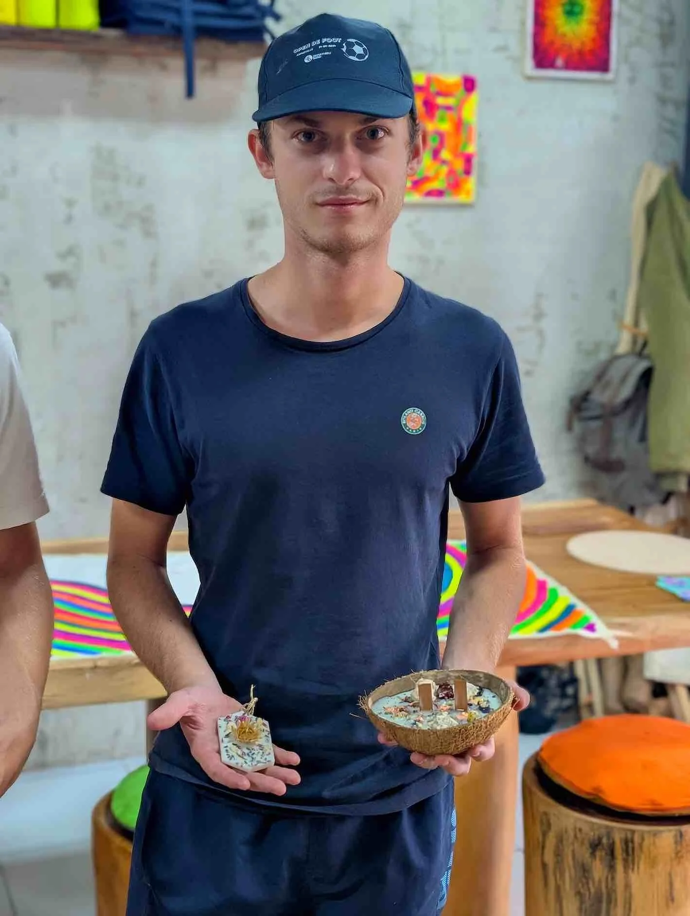 A young man wearing a navy blue cap and t-shirt, holding a coconut shell with a collection of small items inside. The background features colorful artwork and a rustic interior.