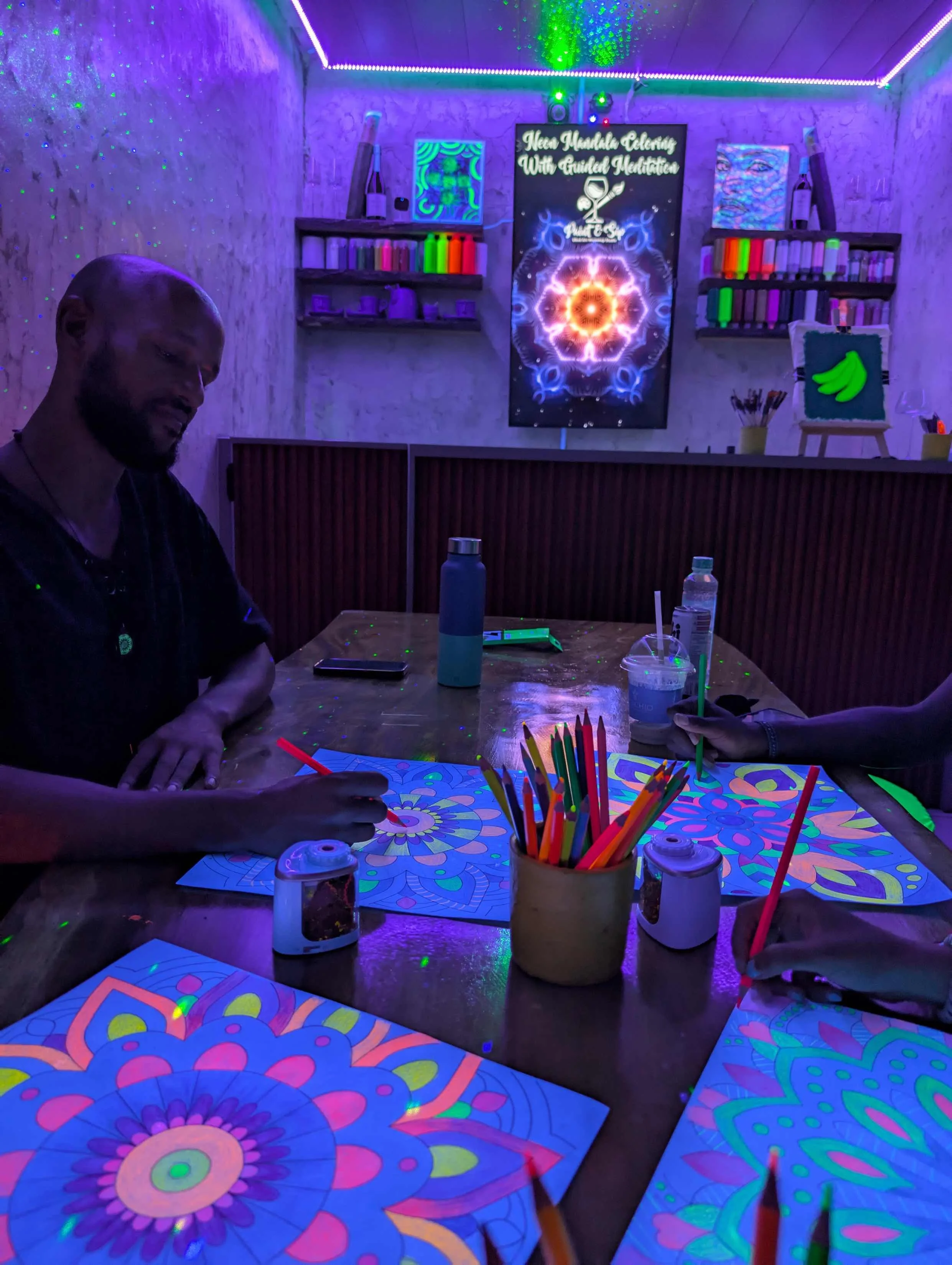 People drawing colorful, glow-in-the-dark mandala art with neon-colored markers in a dimly lit, blacklight room.