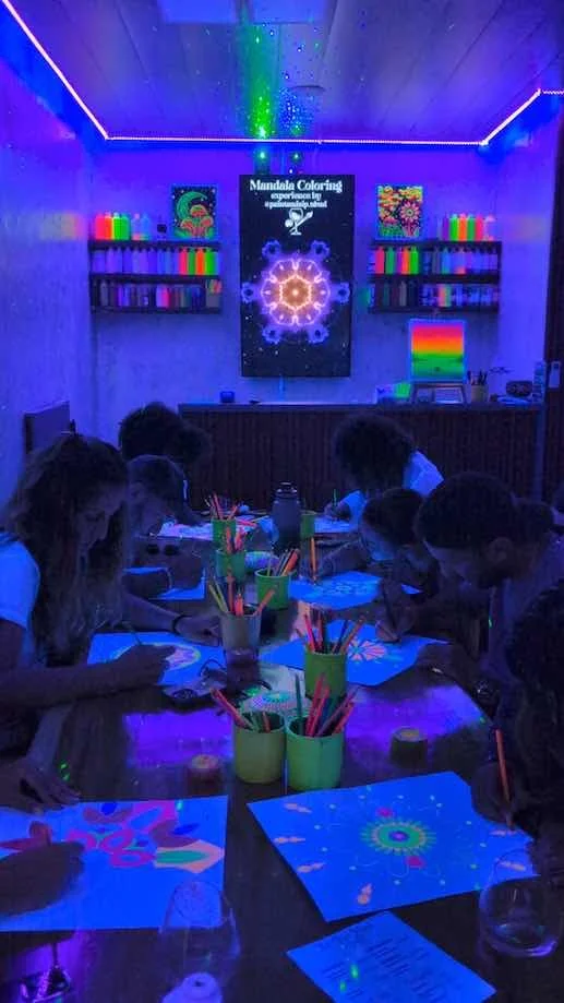 Children participating in a glow-in-the-dark mandala coloring event in a dimly lit room illuminated by UV lights, with colorful art supplies and glowing artworks.