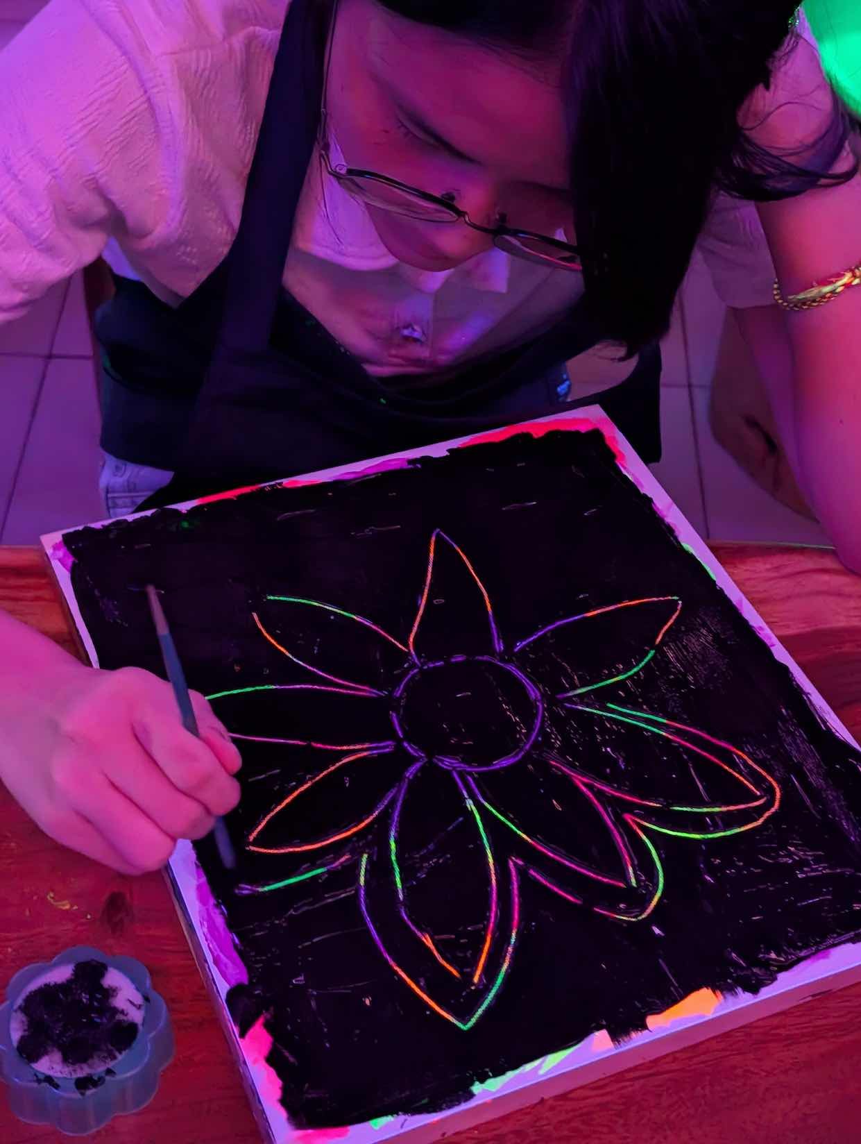 A person with glasses is creating a neon-colored flower drawing on black paper using fluorescent paints under blacklight.