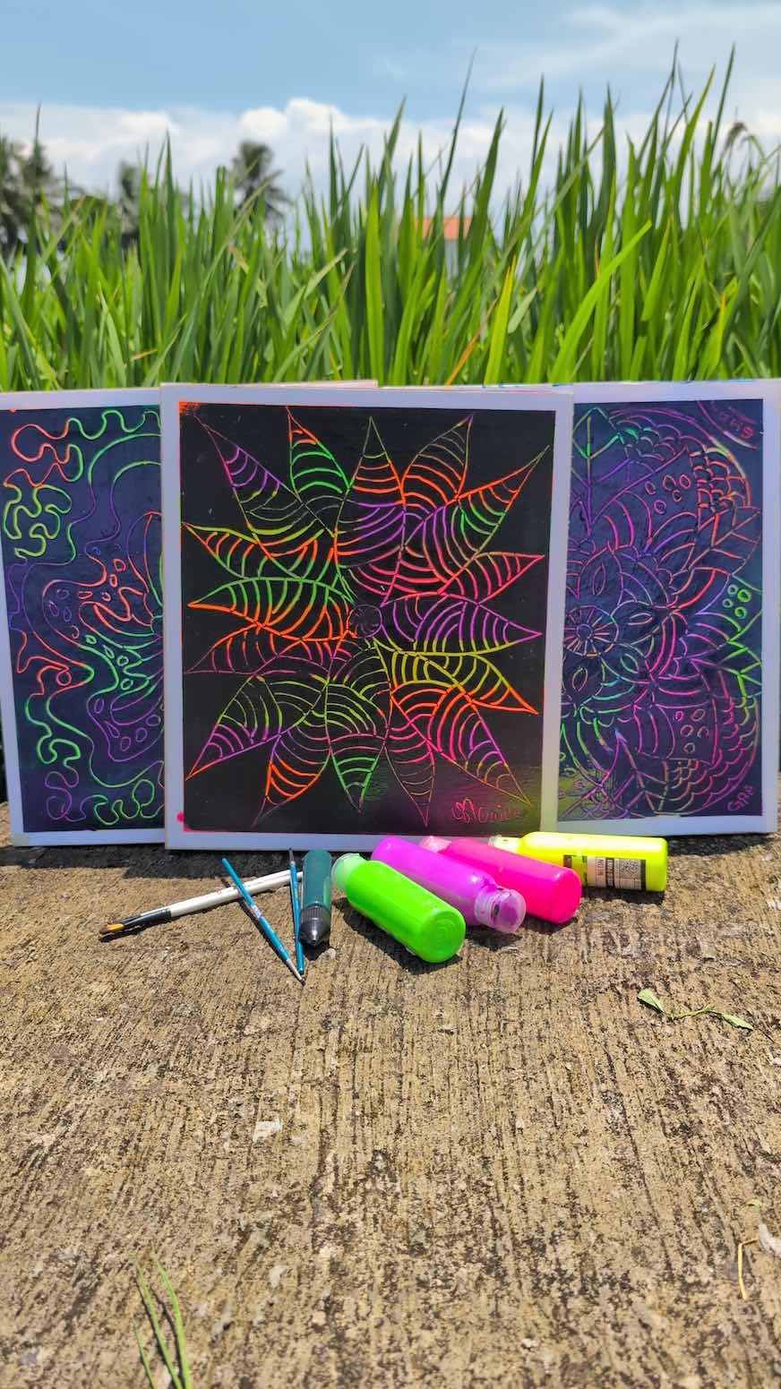 Three neon-colored chalkboard drawings featuring abstract and floral designs, resting on a concrete surface with colorful chalks and pens in front, set against a backdrop of tall green grass and a blue sky with clouds.