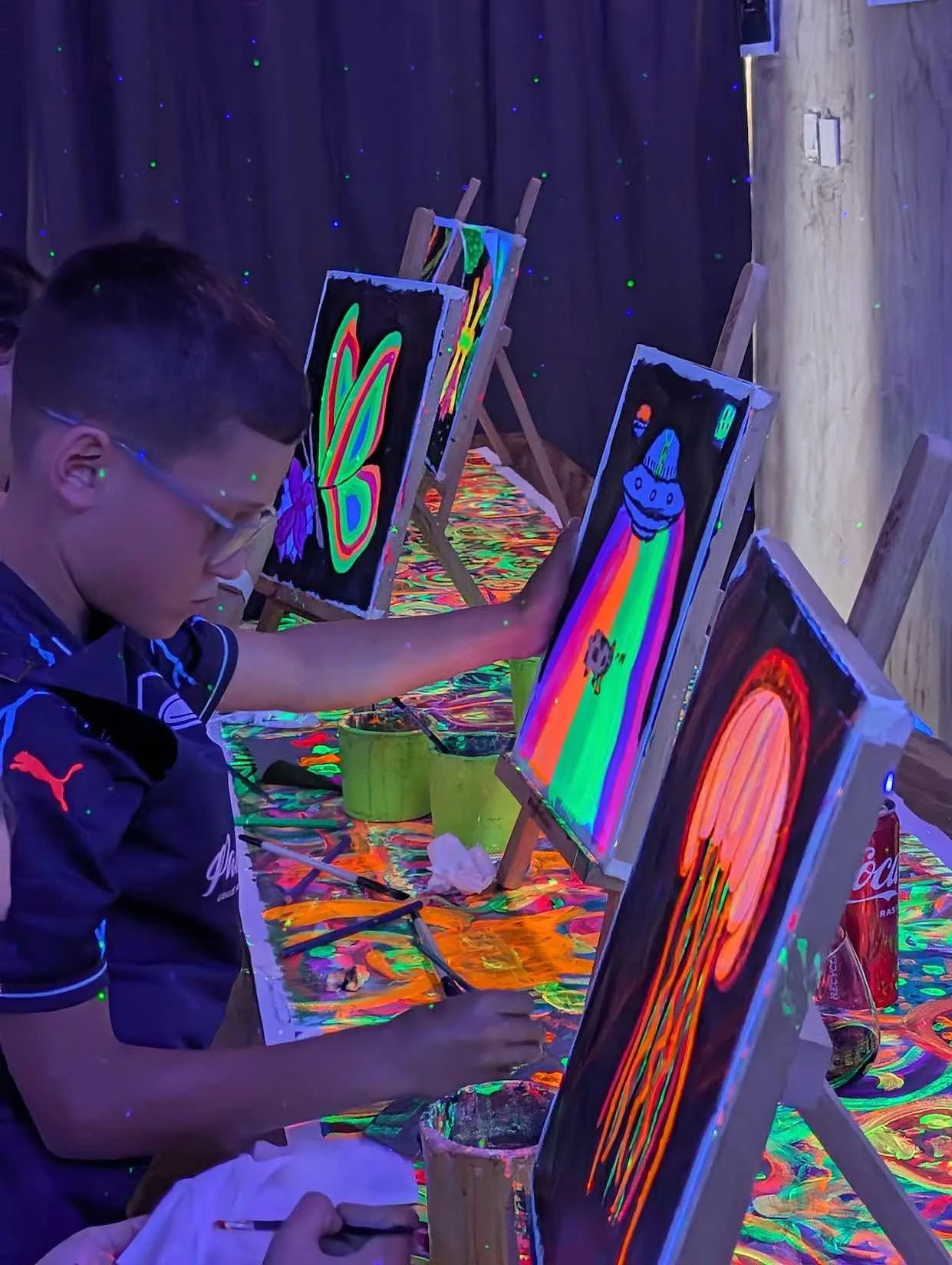 Young boy painting glow-in-the-dark artwork at a painting studio with blacklights, featuring neon colors and abstract designs.