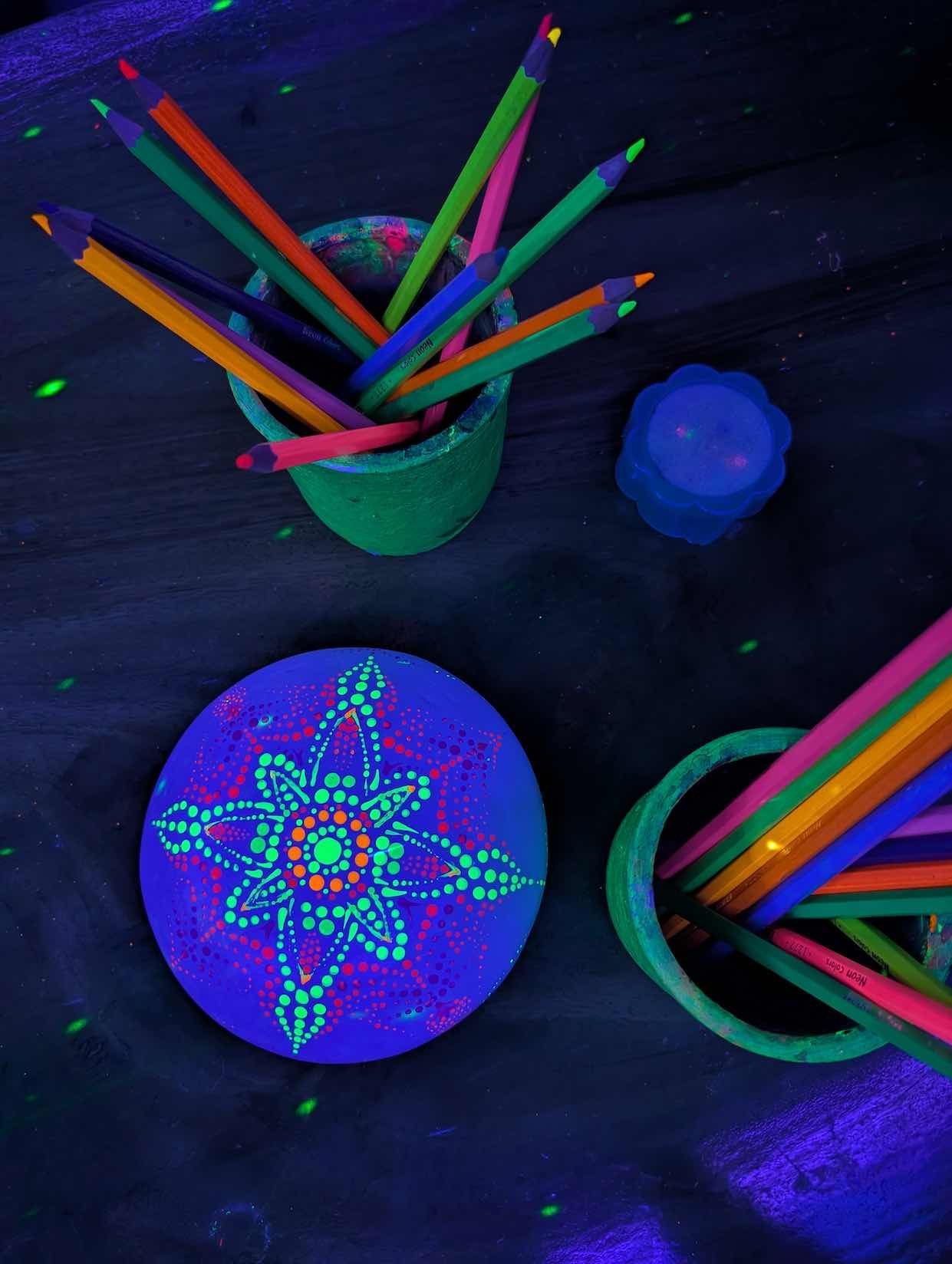 Colorful glow-in-the-dark coloring supplies and decorations on a dark surface, including a bowl of colored pencils, a lantern with a star pattern, and a blue plastic container.