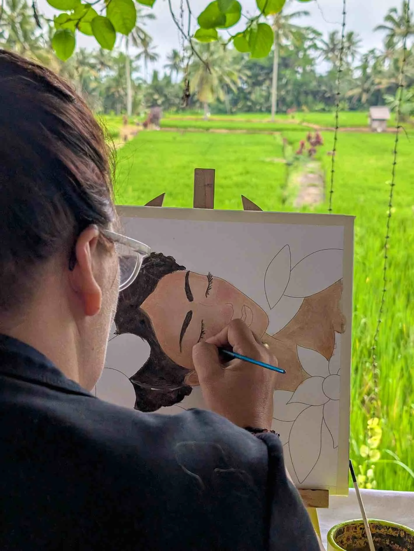 An artist painting a portrait of a woman with closed eyes, dark hair, and long eyelashes, sitting on a chair outdoors with a green rice field and trees in the background.
