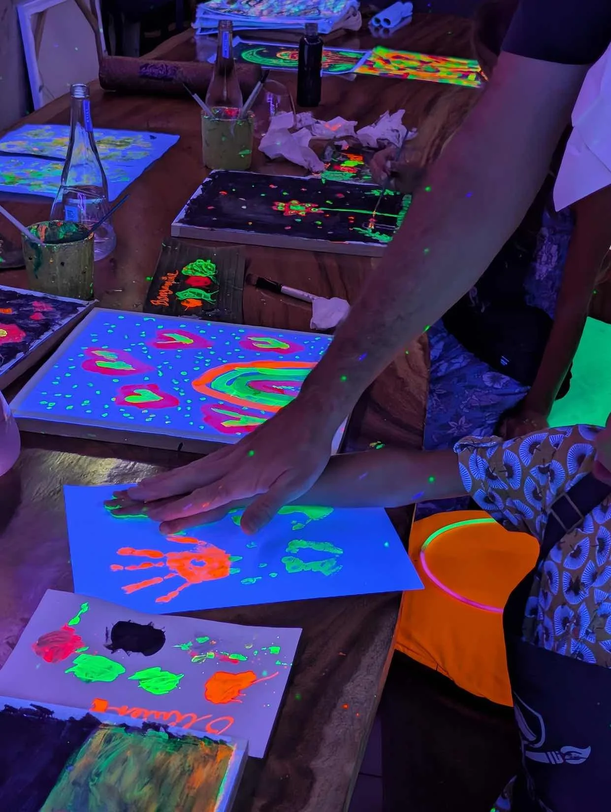 People creating glowing, neon art with black light effects on paper and canvas, using bright paint in a dimly lit room.