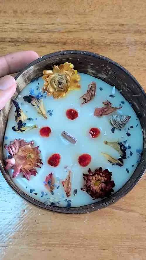 A round candle decorated with dried flowers, small red beads, and tiny shells in a black container.