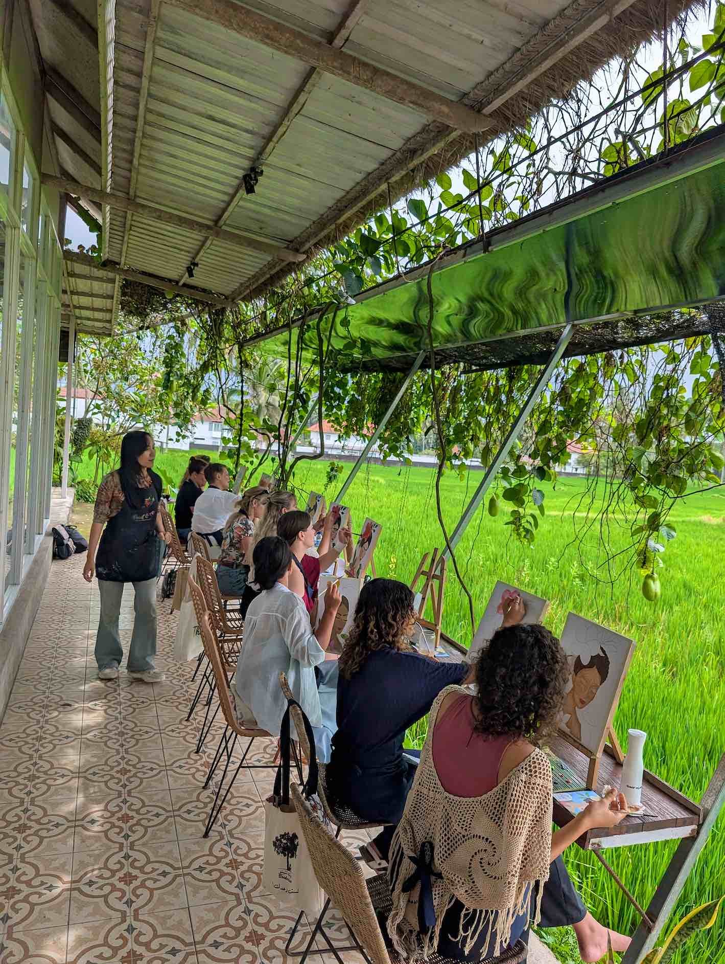 A group of people sitting on chairs outside, painting or drawing on canvases, while overlooking a lush green rice field. A woman stands nearby supervising.