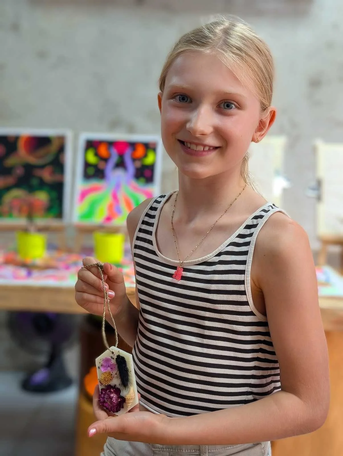 A young girl with blonde hair and a striped tank top smiling while holding a necklace with a decorative pendant, standing in front of colorful artwork and art supplies.