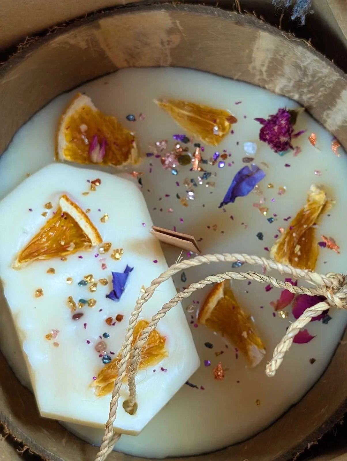 A close-up of a decorated candle with dried orange slices, colorful sprinkles, and flower petals inside a wooden container.