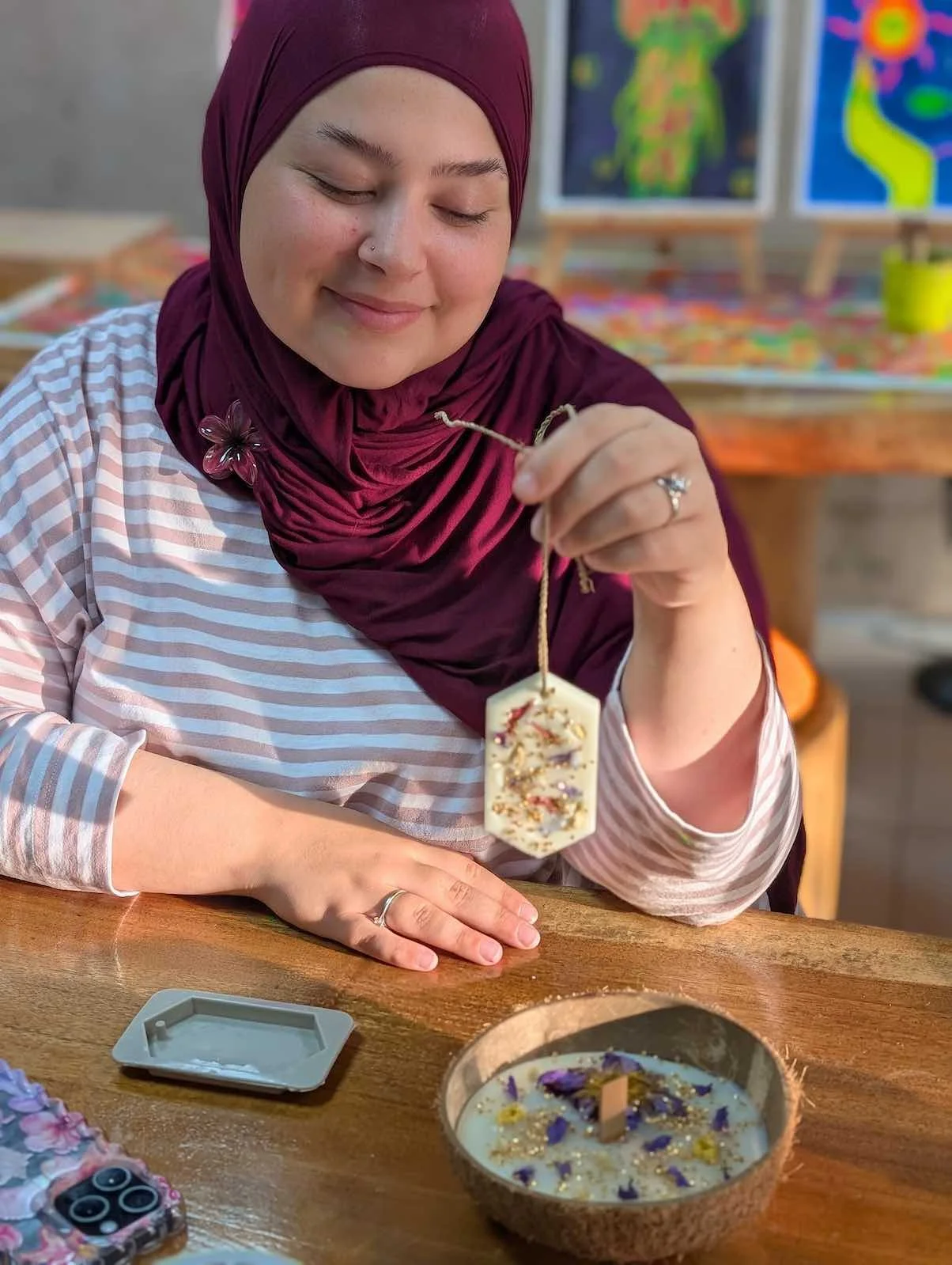 Woman with a maroon hijab and striped shirt holding a decorated soap bar with a string, sitting at a wooden table with a bowl of decorated soap and a phone nearby, in an art studio with colorful artwork in the background.