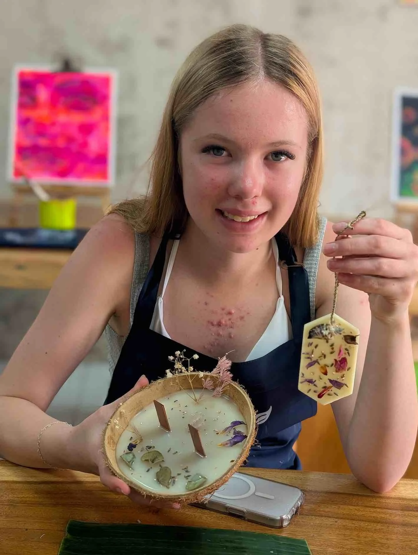 Young woman with red hair holding a decorative soap shaped like a dish in her hand and a matching soap in a bowl on a wooden table, with colorful artwork in the background.