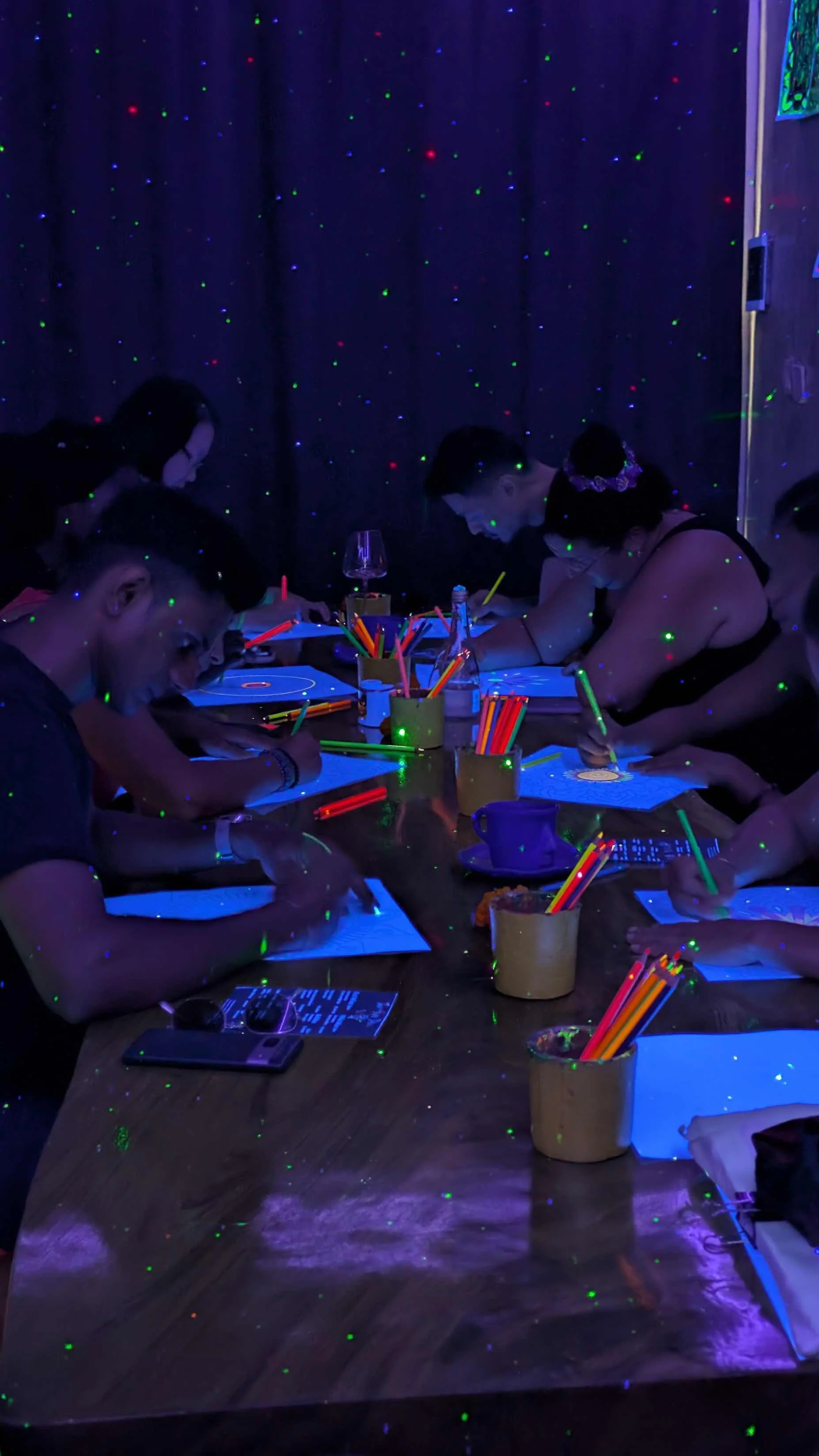 Group of people sitting at a table under black light, drawing with neon-colored markers, with glow-in-the-dark dots on the wall and table.