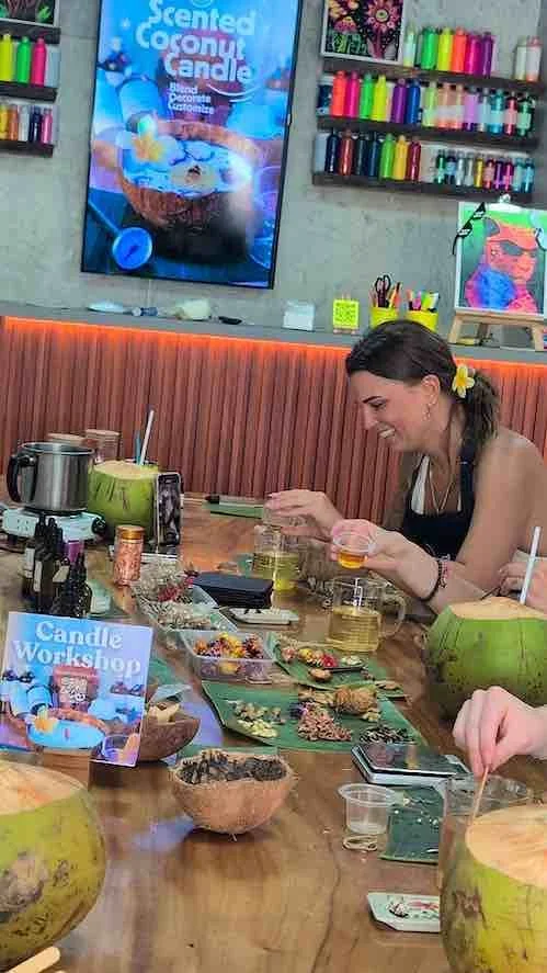 A woman at a candle workshop, sitting at a table with coconut shells, candles, and crafting supplies, in a store with colorful shelves of candle wax and a sign that reads "Candle Workshop."