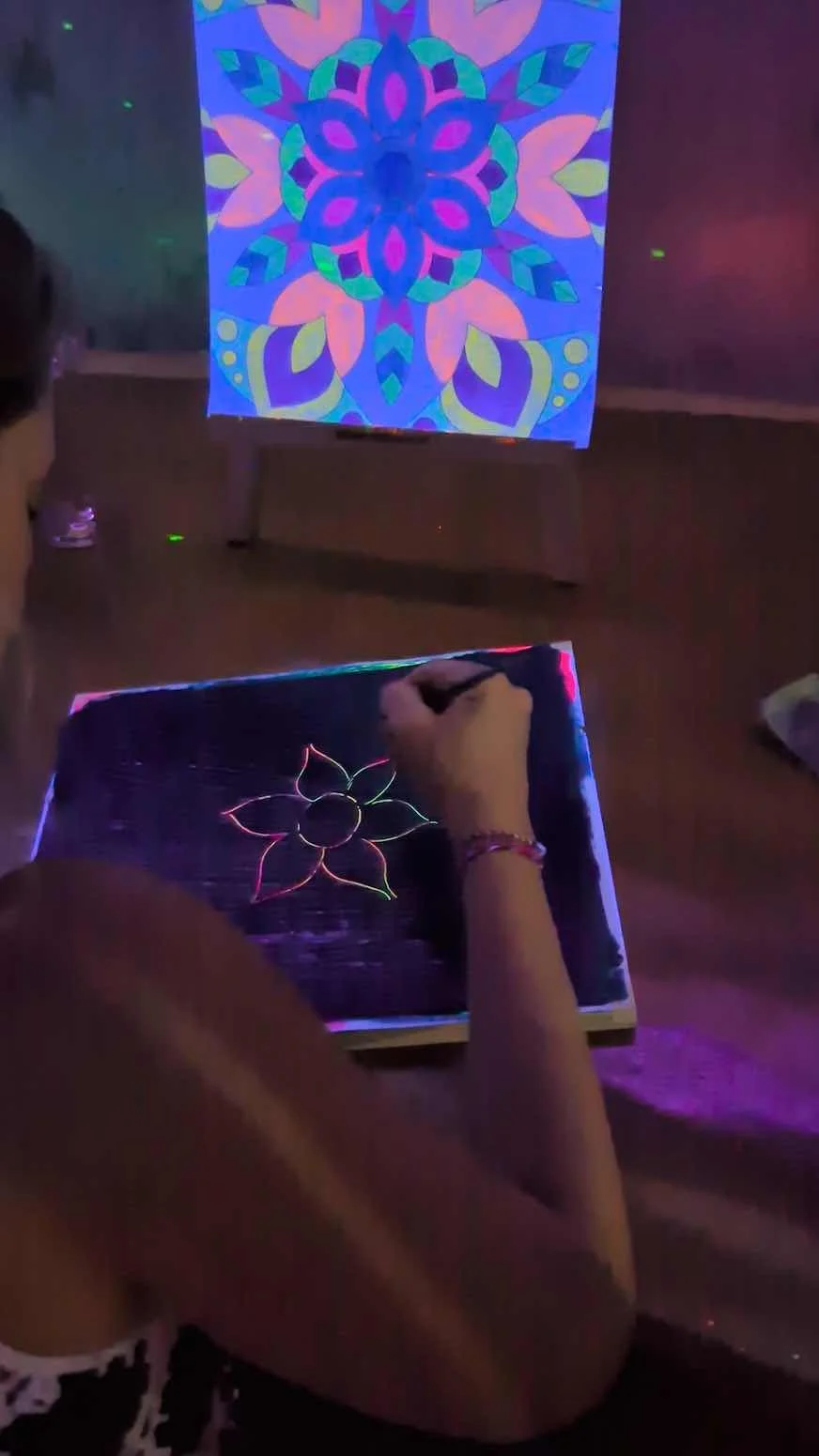 Person drawing a glow-in-the-dark flower on a black canvas, with a colorful mandala pattern projected on the wall in the background.