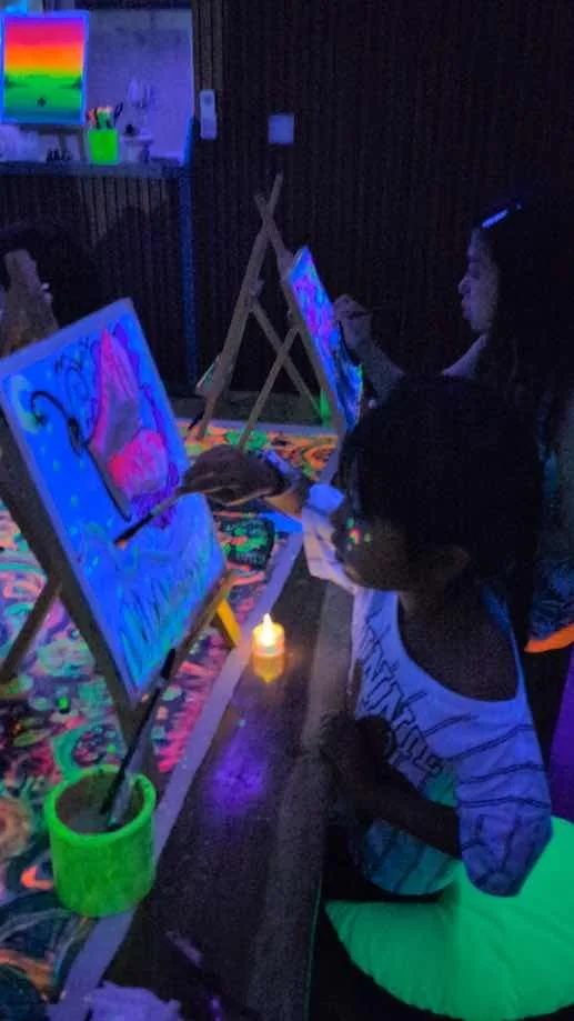 Children painting on canvases in a dark room illuminated by blacklight, with neon colors and a small candle on the floor.