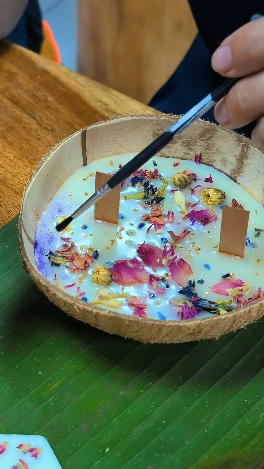 A hand holding a paintbrush decorating a round candle with dried flowers, gold beads, and small wooden sticks inside a wooden bowl.