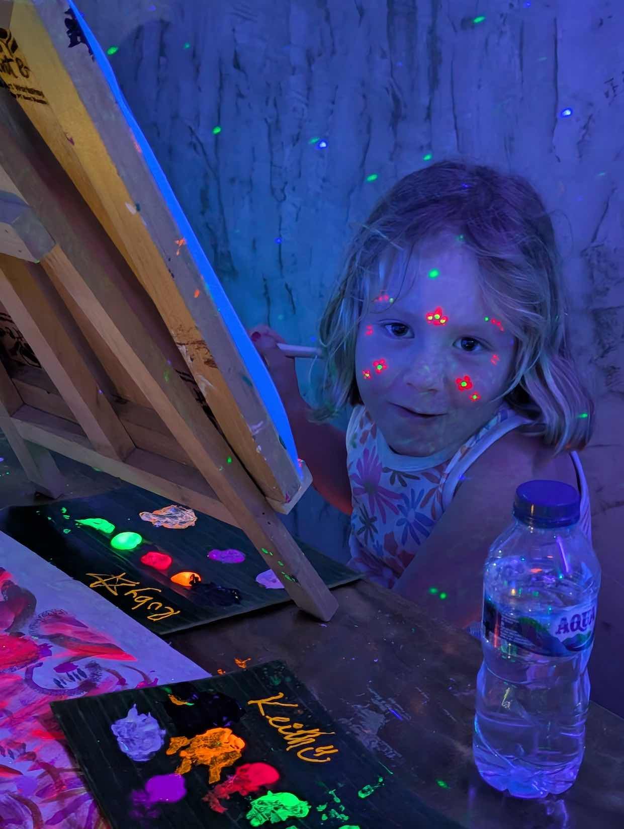 A young girl with face paint under blacklight, painting on an easel with colorful paints, in a dimly lit room with blacklight effects.