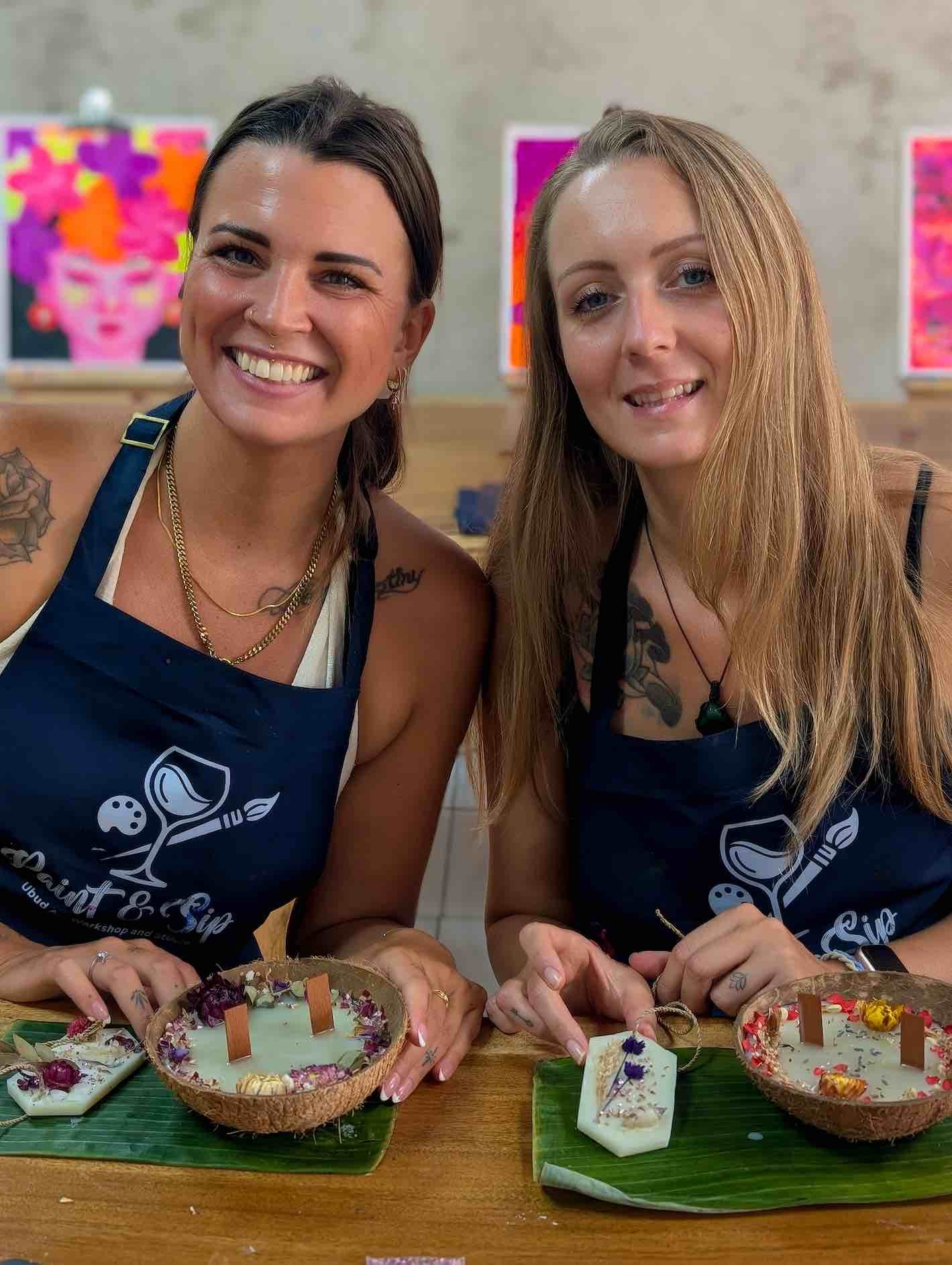 Two women wearing matching aprons sit at a table, showcasing jewelry and handcrafted art pieces displayed in coconut shells and on plates, with colorful artwork hanging in the background.