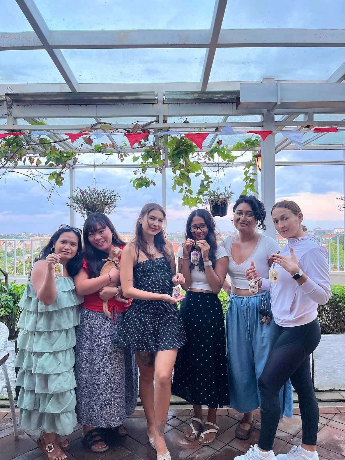 A group of seven women standing together on a rooftop terrace, holding refreshments, with a city skyline in the background and a decorated pergola overhead.