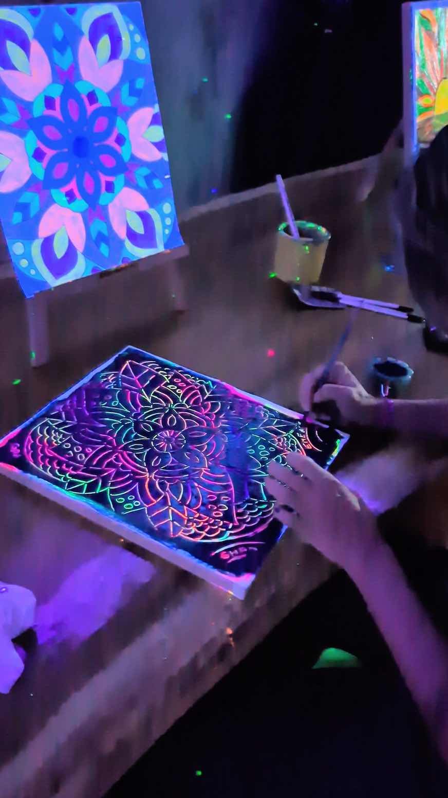Person drawing a colorful, glow-in-the-dark mandala pattern on a tablet with neon lights reflecting in a dark room, with abstract neon artwork in the background.