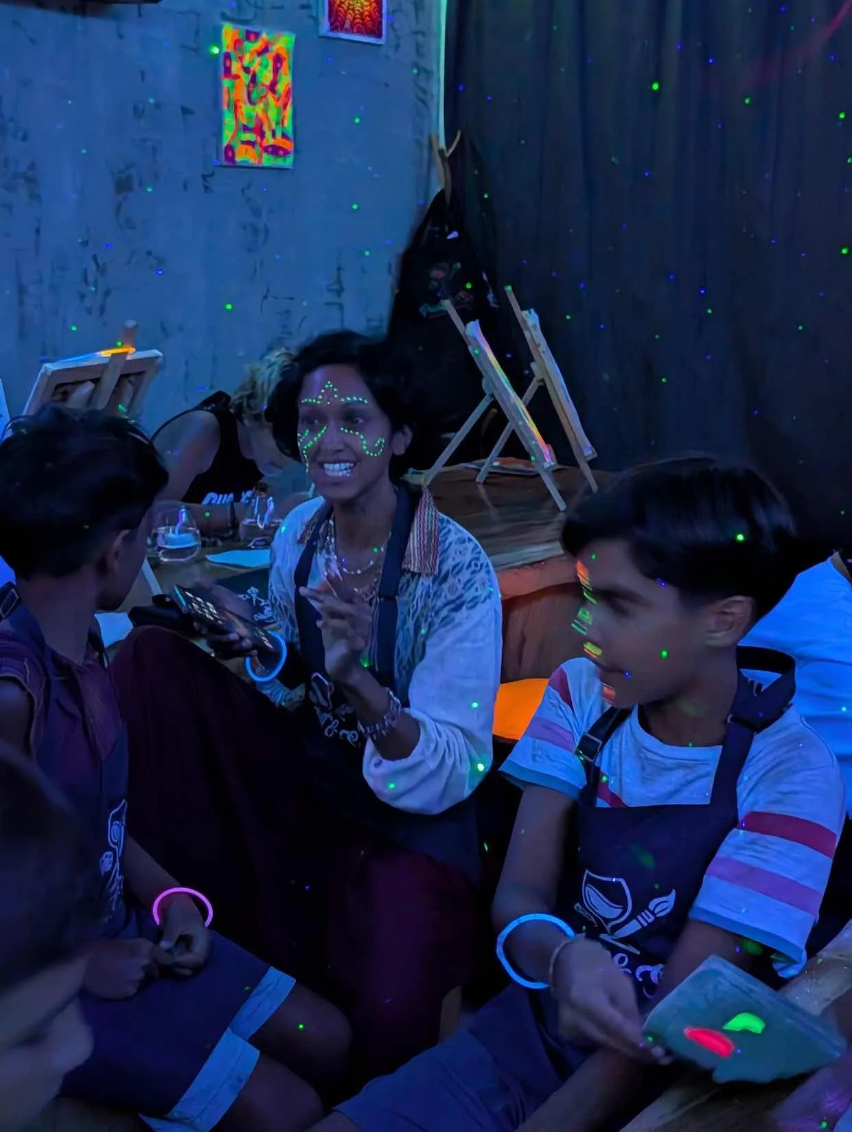 Group of people in a dark room with blacklight effects, the woman in the center has face paint that glows and is smiling and talking, while the others are seated around her with glowing accessories, there are artwork and miniature easels in the backg