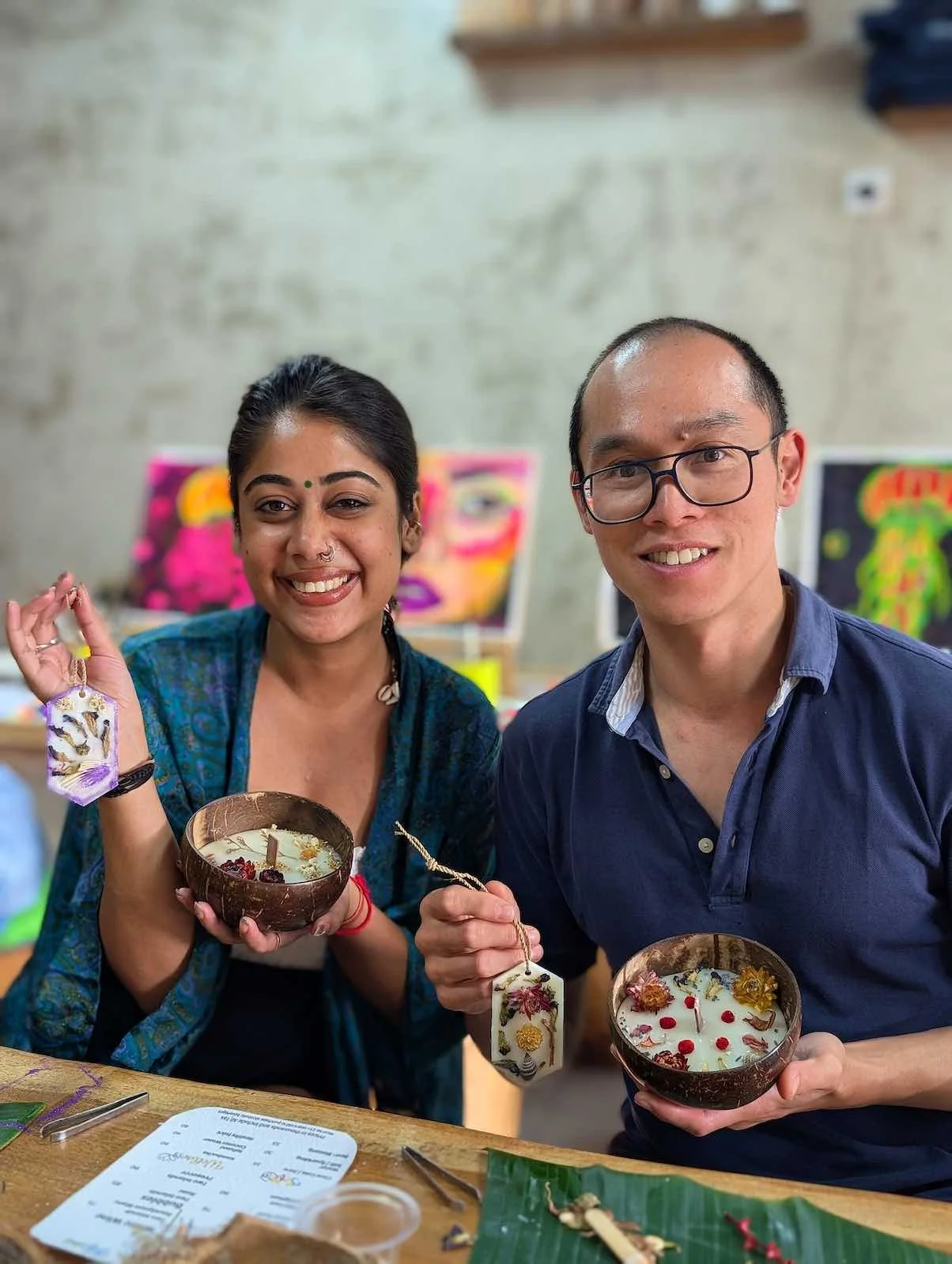 Two people smiling while holding decorated bowls in an art studio, with colorful artwork in the background.