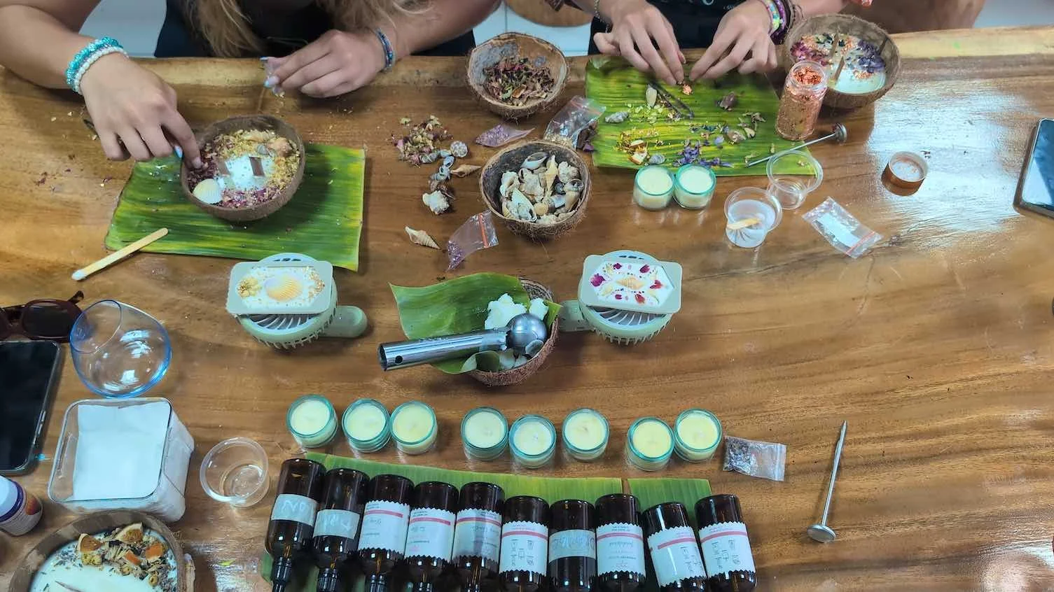 A table with various candles, essential oils, shells, and tools used for making aromatherapy or herbal products. Hands of people are working with natural ingredients on banana leaves.