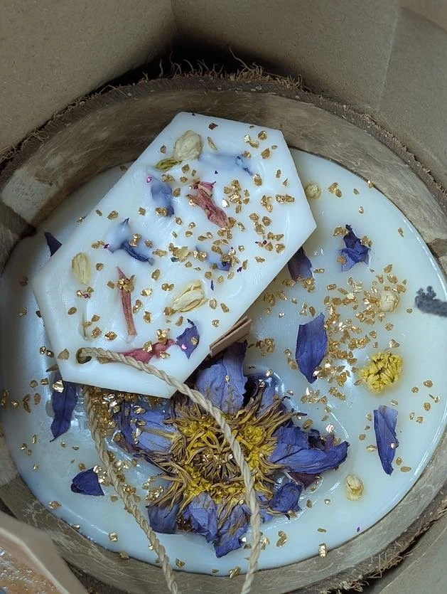 A white candle decorated with purple flower petals and gold flakes, placed inside a wooden container filled with wax and dried flower petals.